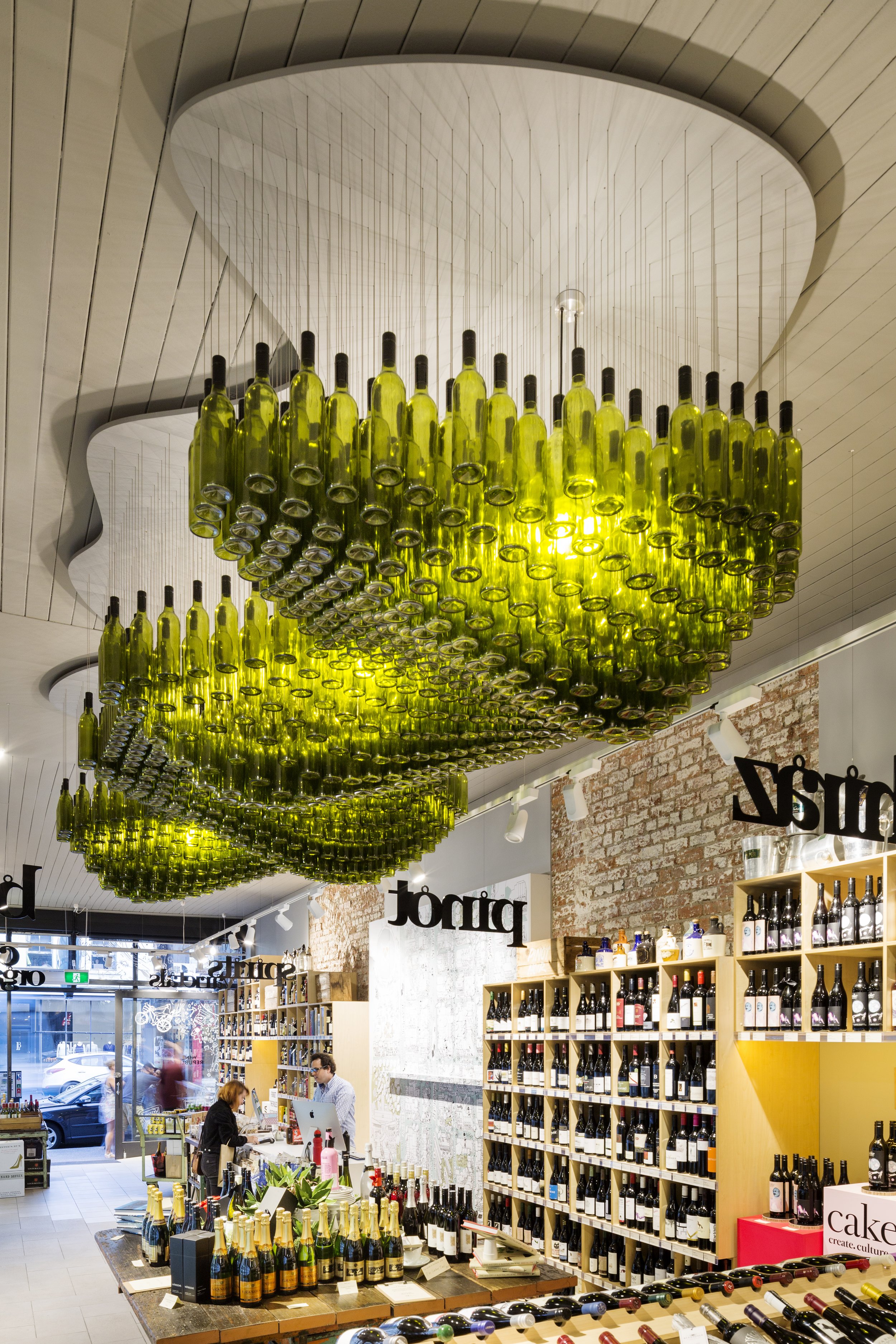 Inside a wine store featuring a ceiling installation of upside-down wine bottles illuminated with yellow light, shelves filled with wine bottles, and people shopping and working at the counter.