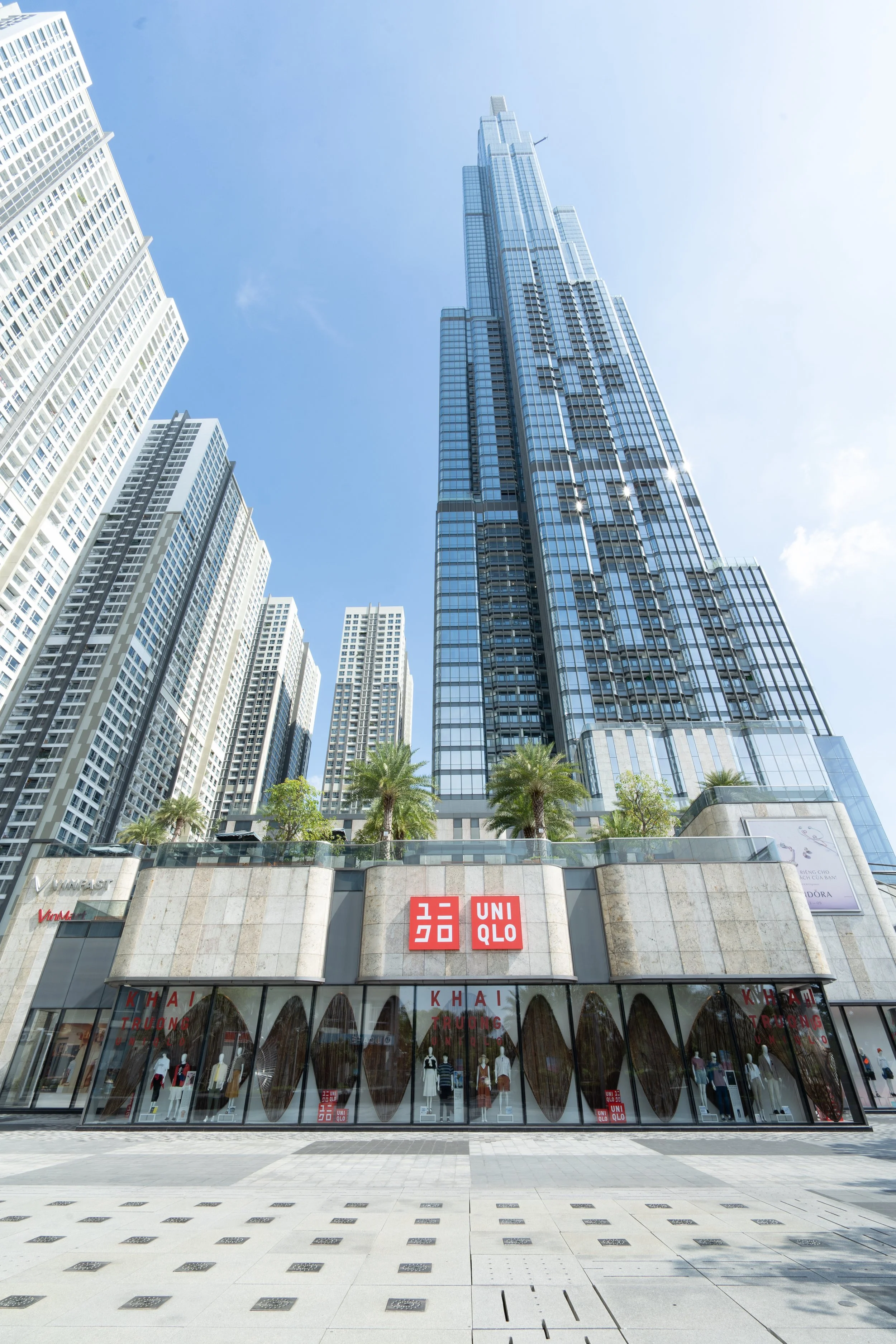 A high-rise glass skyscraper with a smaller building at its base, featuring store signs for Uniqlo and other brands at street level. The sky is blue and clear with some clouds.
