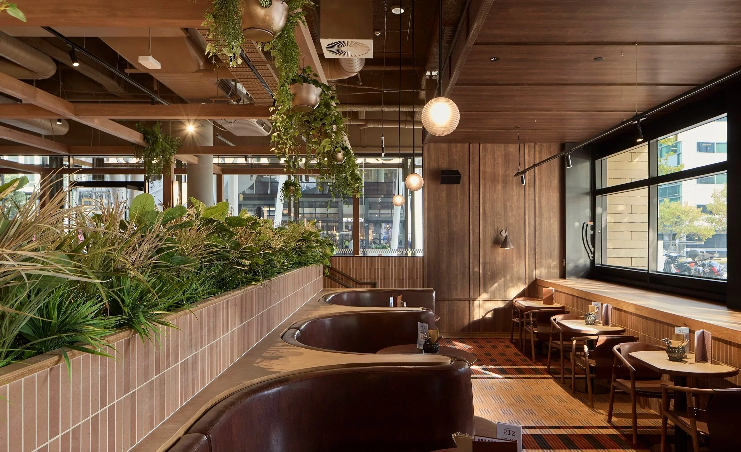 Interior view of a modern restaurant with wooden paneling, hanging plants, spherical pendant lights, and large windows showing a cityscape outside.