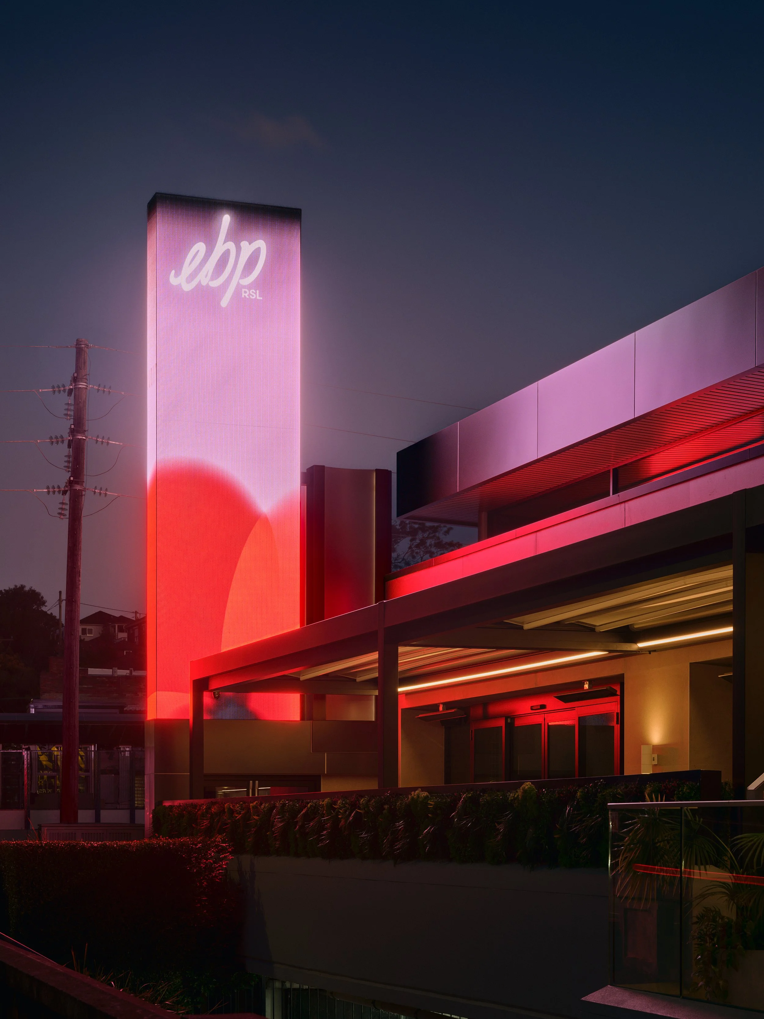 Nighttime exterior view of a modern building with a tall illuminated sign displaying the logo 'ebp RSL' in white cursive on a pinkish-red background. The building has sleek architectural design with red accent lighting under the overhang and surrounding greenery in the foreground.