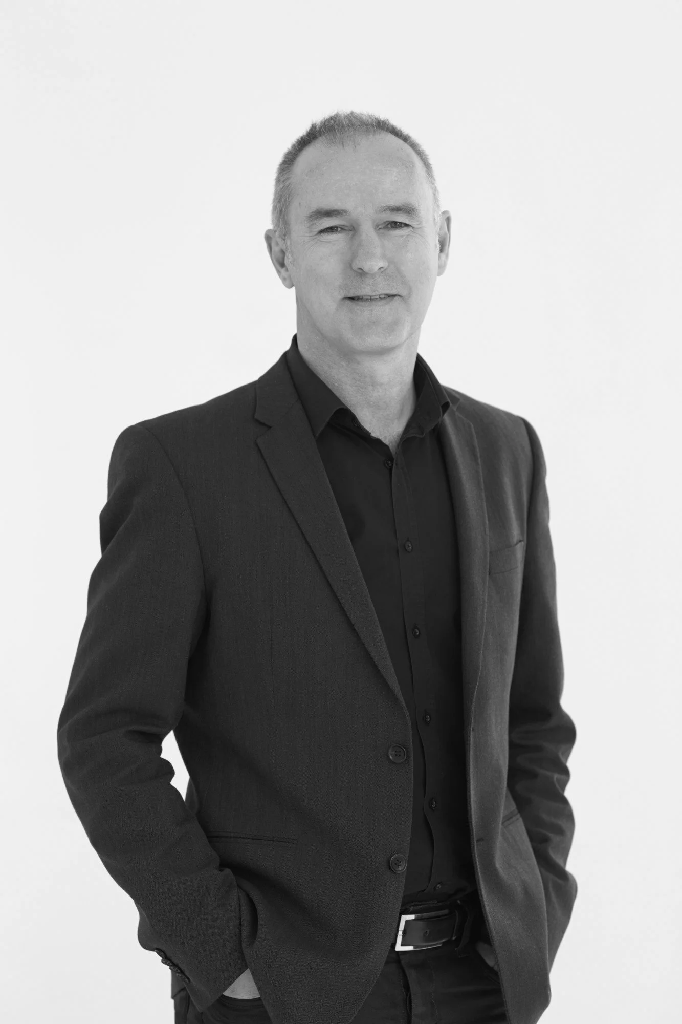A portrait of a middle-aged man with short hair, wearing a dark suit, standing against a plain white background.