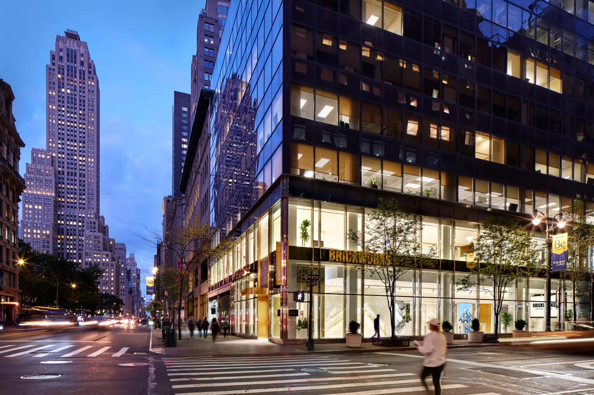 City street scene at dusk with modern glass office building on the right, historic skyscrapers on the left, and a busy crosswalk with people walking across.