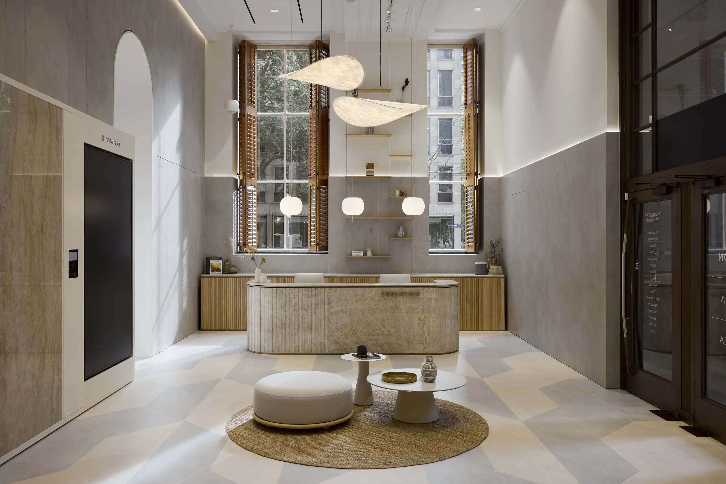 Modern lobby or reception area with large windows, wooden blinds, circular pendant lights, and a marble reception desk with decorative vases and books. There are two small round tables with a cushion in front, and a rounded rug underneath. The space is well-lit with natural and artificial light.