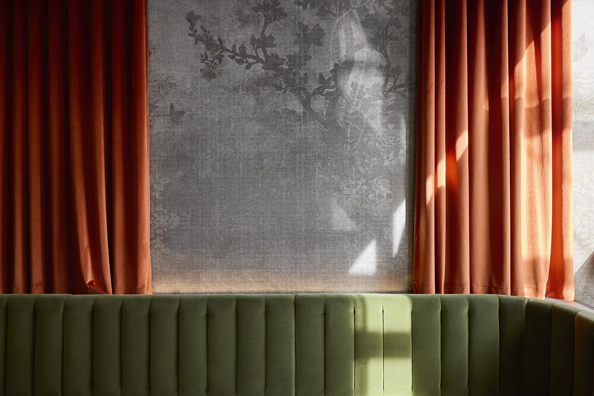 Green upholstered bench in front of a wall with gray textured wallpaper featuring a subtle tree branch pattern, framed by orange curtains on both sides.