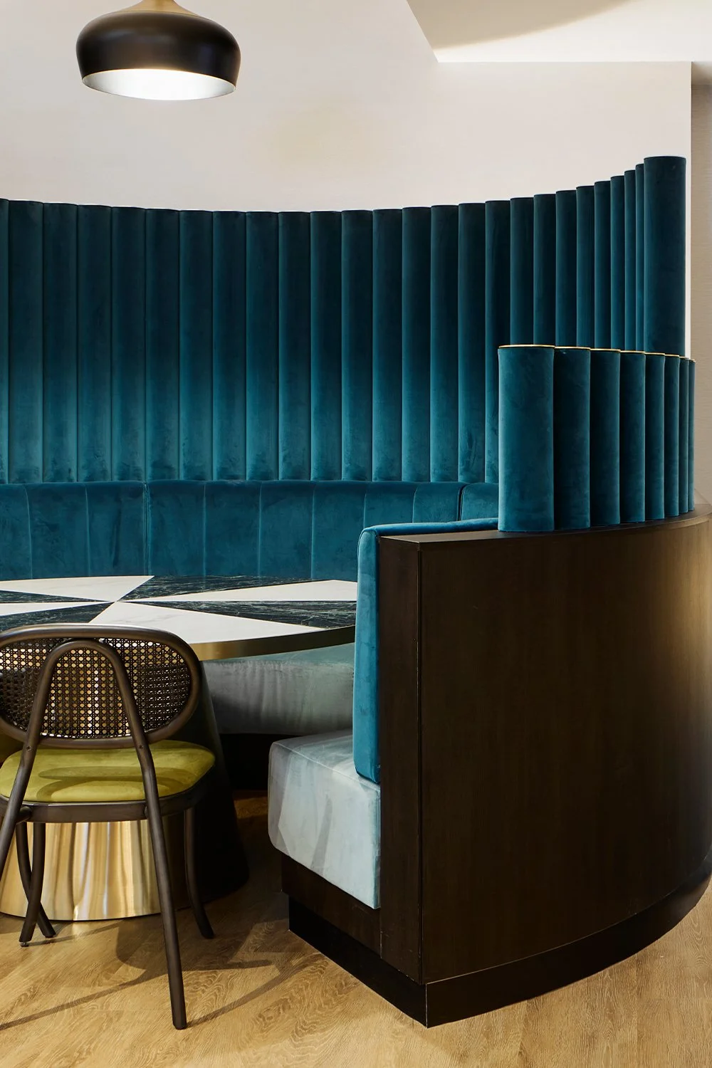 Interior of a modern lounge with a curved, deep teal velvet booth, a black and white geometric table, a rattan chair with a green cushion, and a black pendant light.