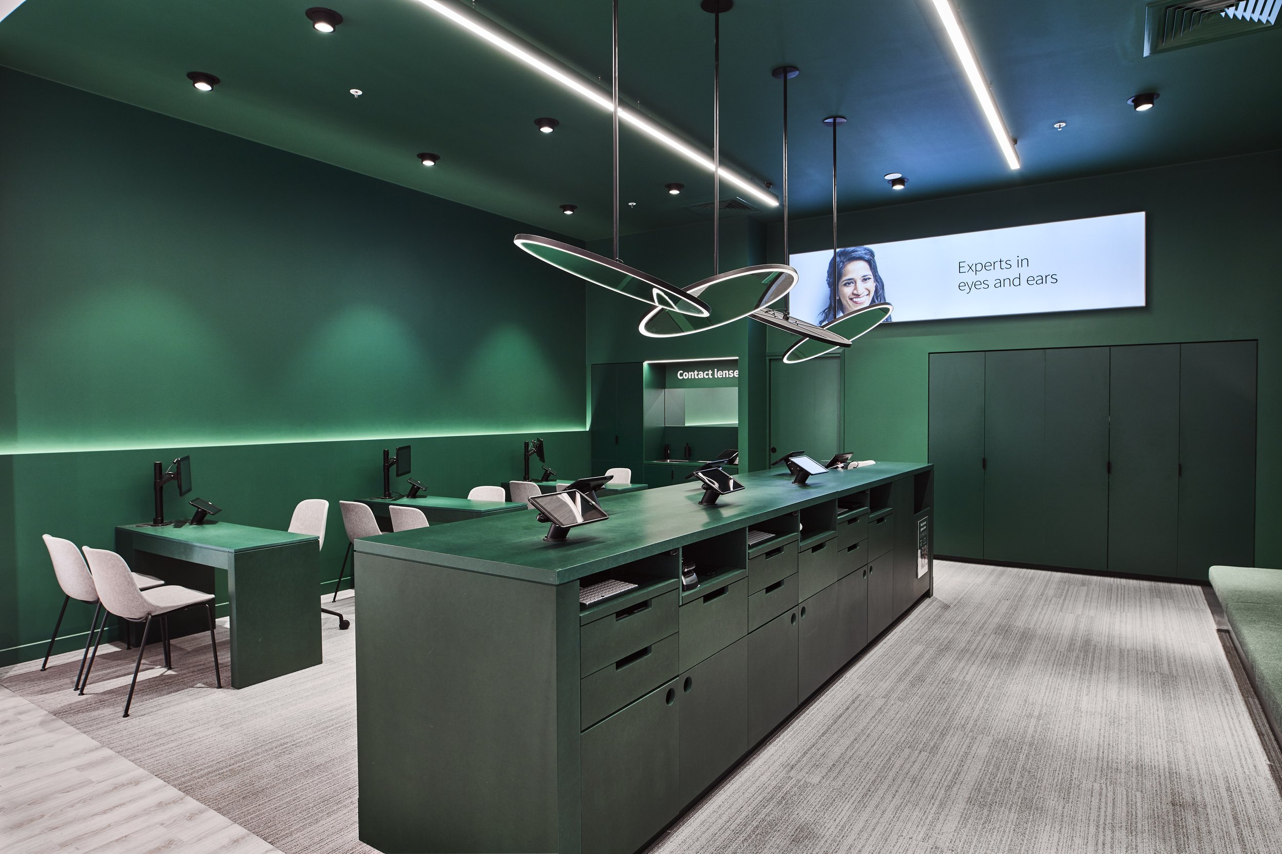 Interior of an eye care clinic with green walls, a long green counter with multiple workstations, white and pink chairs, a large digital screen displaying a woman's face and the text "Experts in eyes and ears," and hanging modern light fixtures.