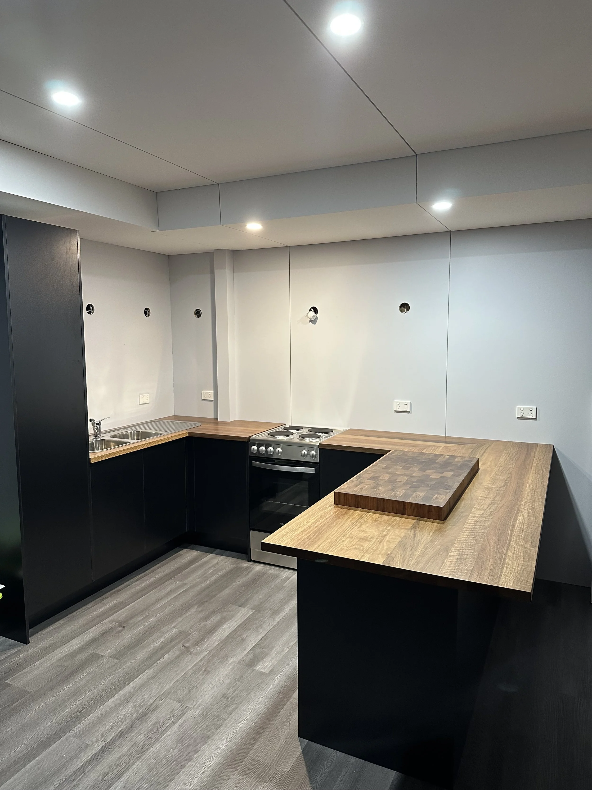 Empty kitchen with black cabinets, wooden countertops, a stove, a sink, and a small wooden cutting board on the counter. The walls are white, and the ceiling has recessed lighting.