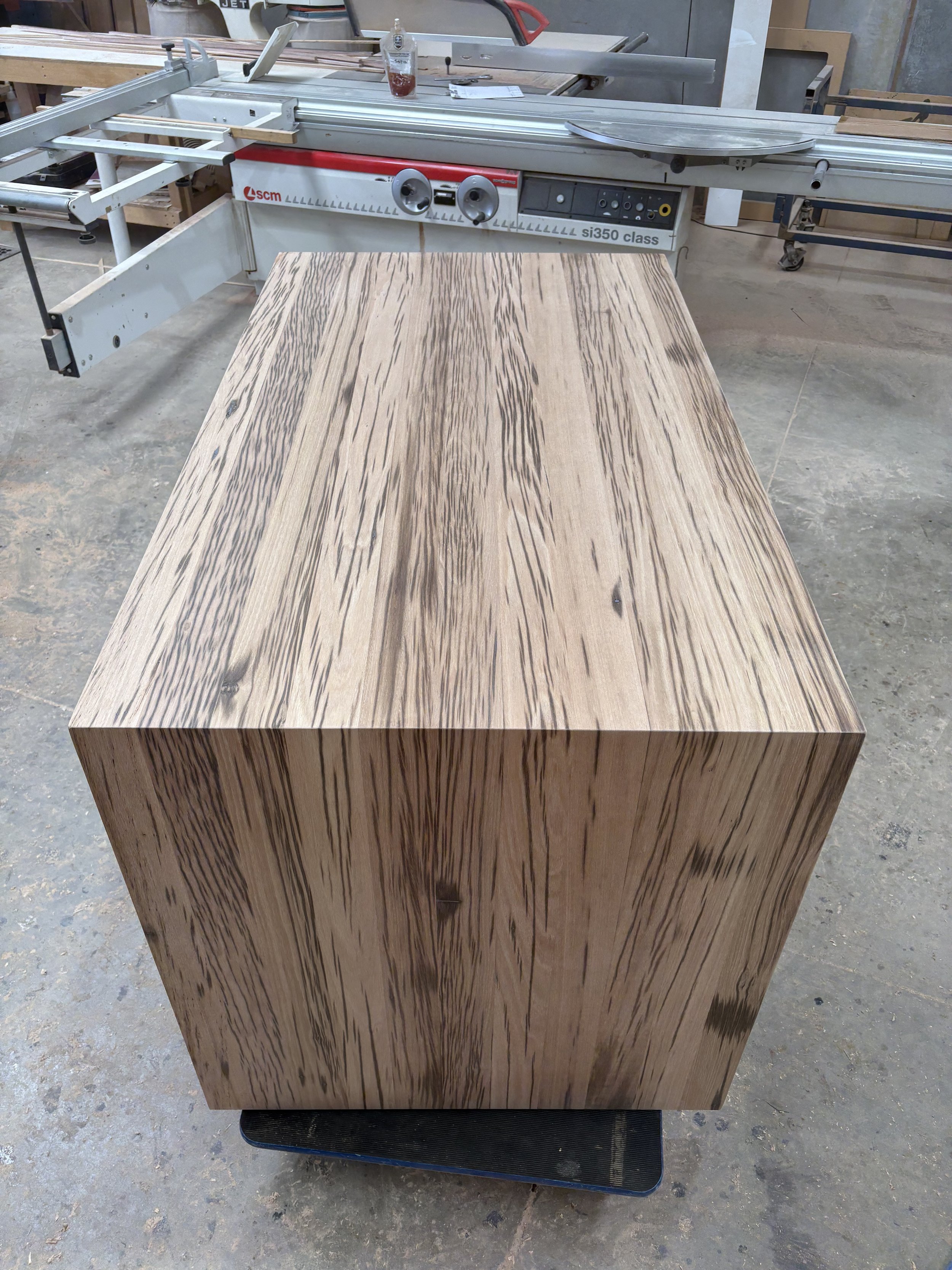A large rectangular piece of wood with a distinctive grain pattern lay on a platform in a woodworking shop. A table saw is visible in the background with various tools and supplies.