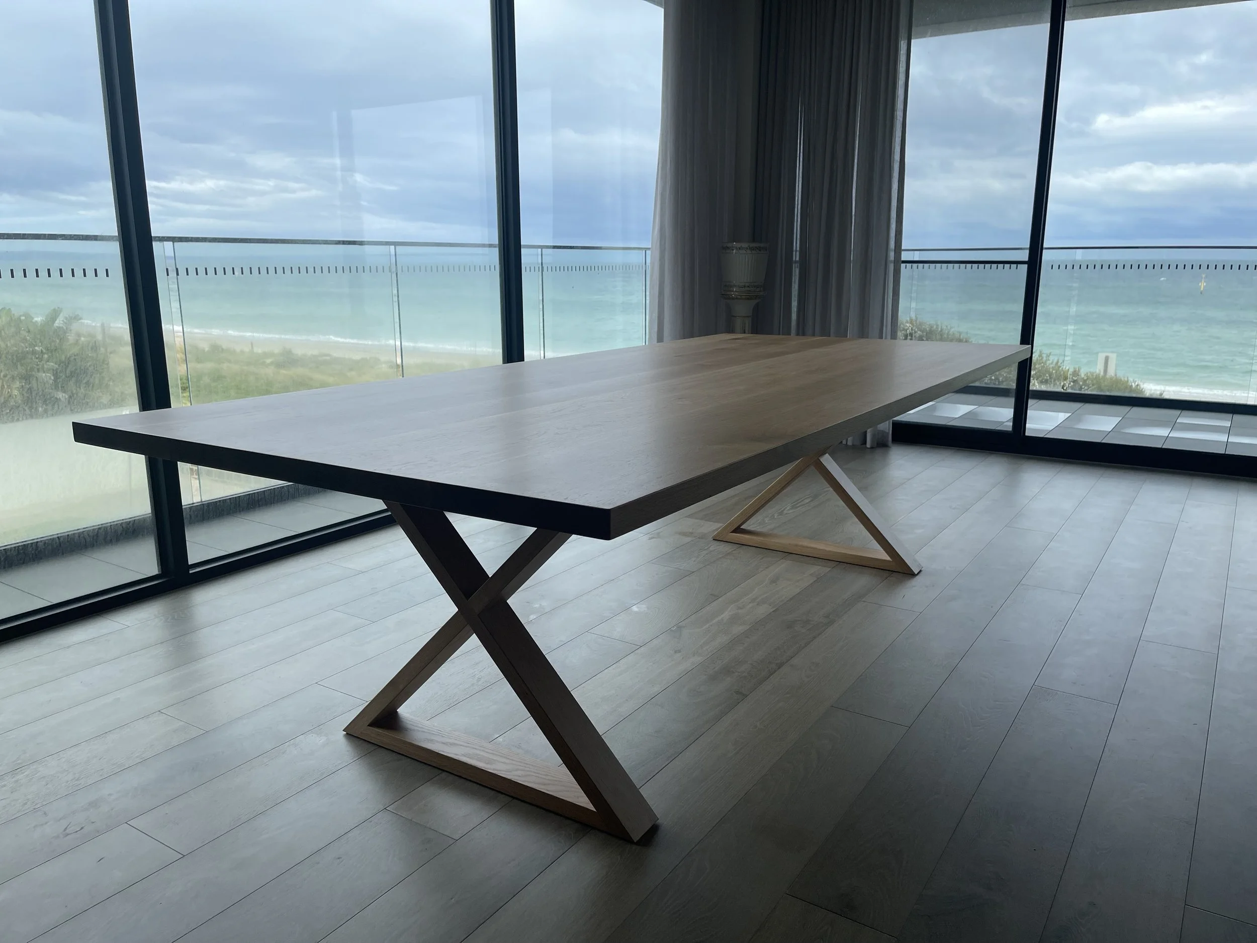 Modern wooden dining table with a unique geometric base in a room with floor-to-ceiling windows overlooking an ocean view.