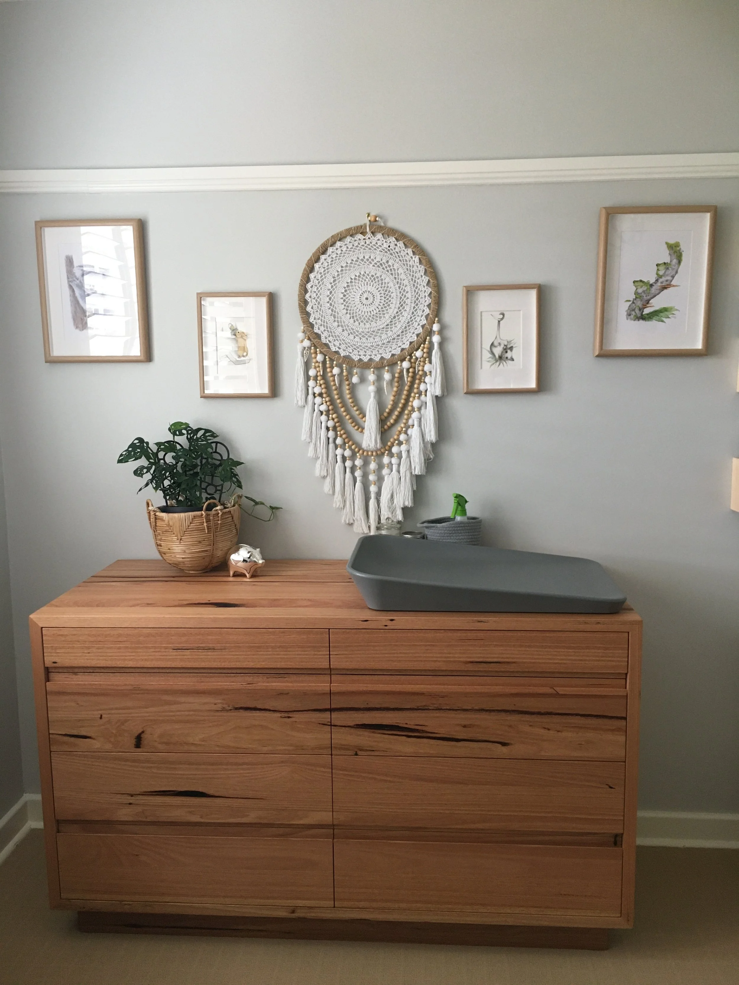 A wooden dresser with six drawers, topped with a gray changing pad, and decorated with a potted plant, a small decorative object, and a spray bottle. Behind it is a wall decorated with framed artwork and a large dreamcatcher with beads and tassels.