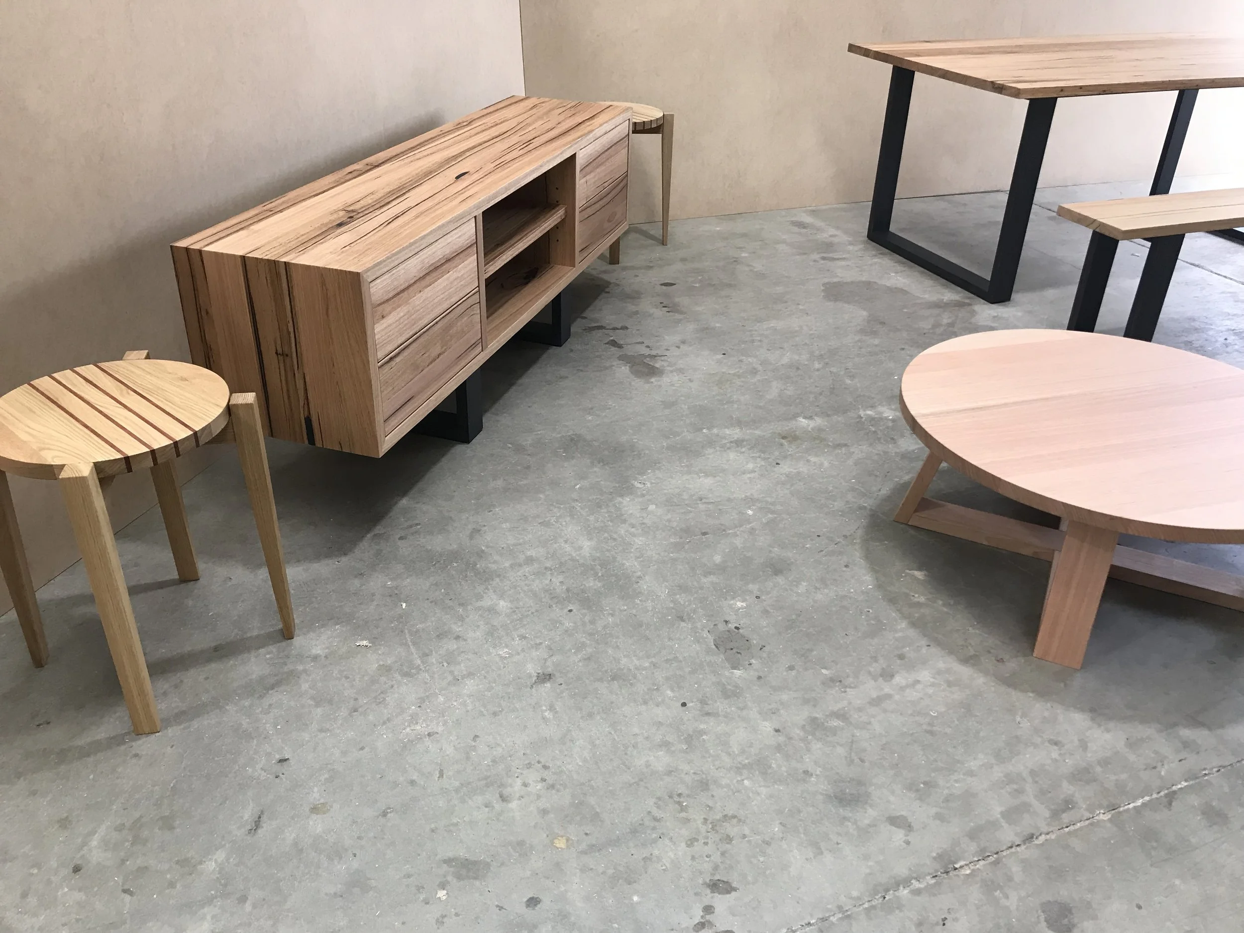 A collection of wooden furniture including a round table, a rectangular table, a small side table, and a long, low cabinet with open shelving, all placed on a concrete floor against a beige wall.