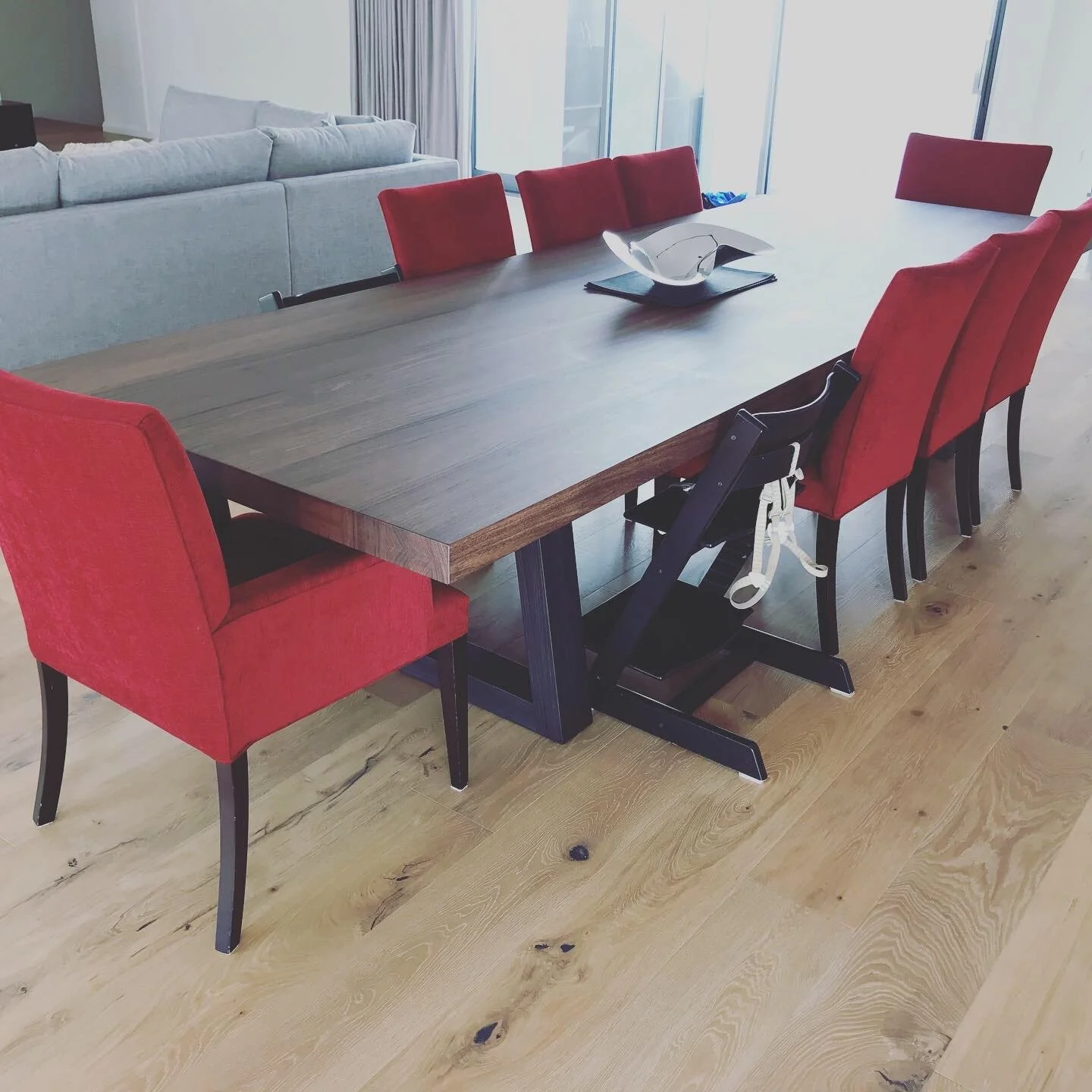 A dining table with red upholstered chairs in a modern living room with hardwood floors and a gray sofa in the background.