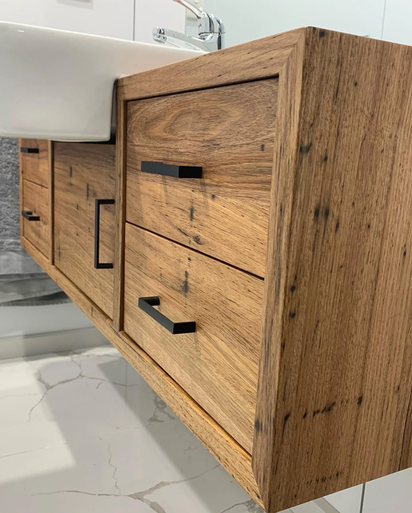 A wooden bathroom vanity with black handles, next to a white sink and chrome faucet, on a marble floor.