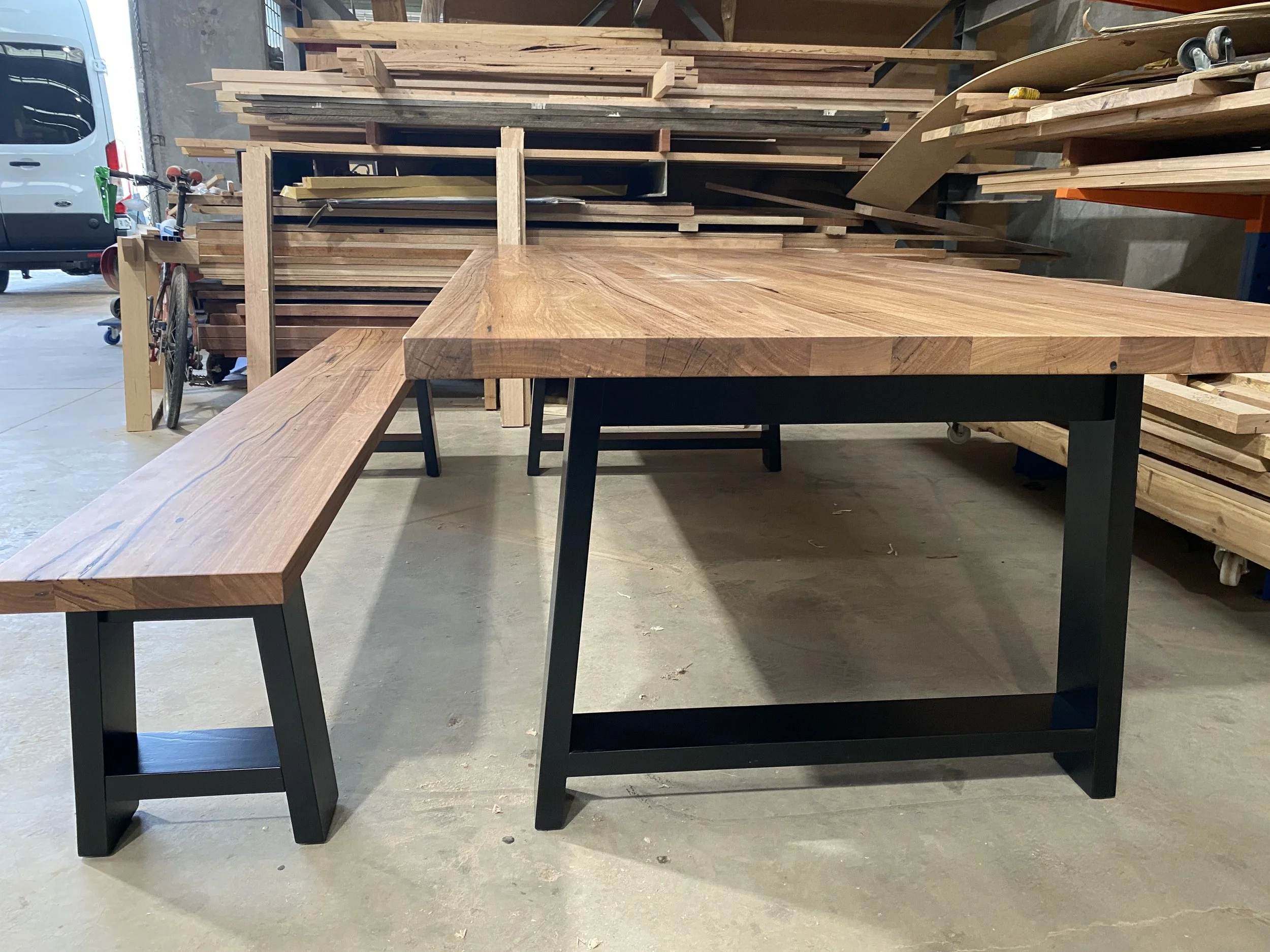 A wooden table with black metal legs and a matching wooden bench with black legs, set in a woodworking workshop with stacks of wood and a bicycle in the background.