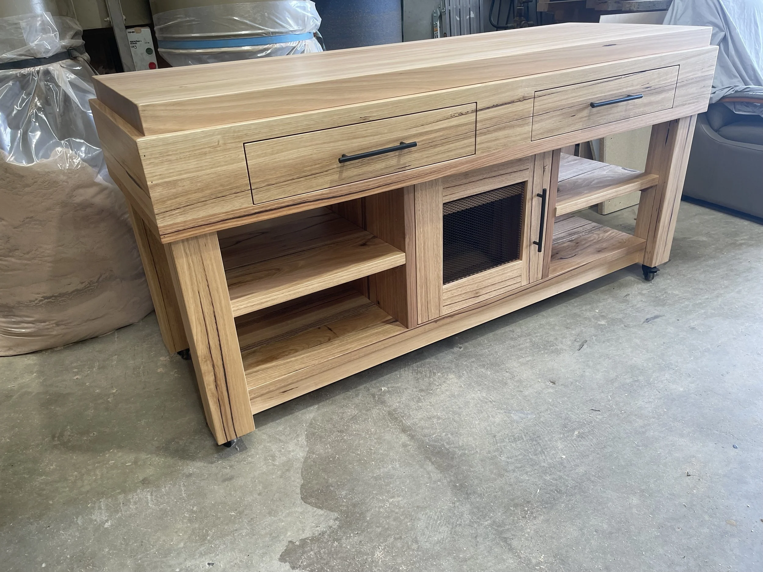 A wooden sideboard or console table with two drawers, a glass-paneled cabinet door, open shelving on both sides, and black handles, situated on a concrete floor in a storage or workshop area.