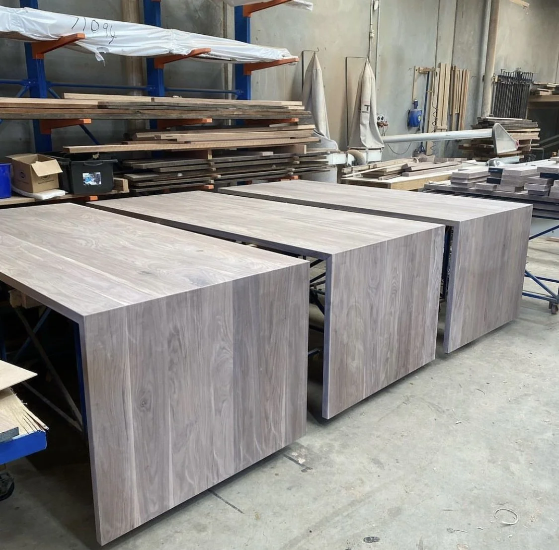 Three wooden tables in a woodworking workshop, with shelves and stacks of wood planks in the background.