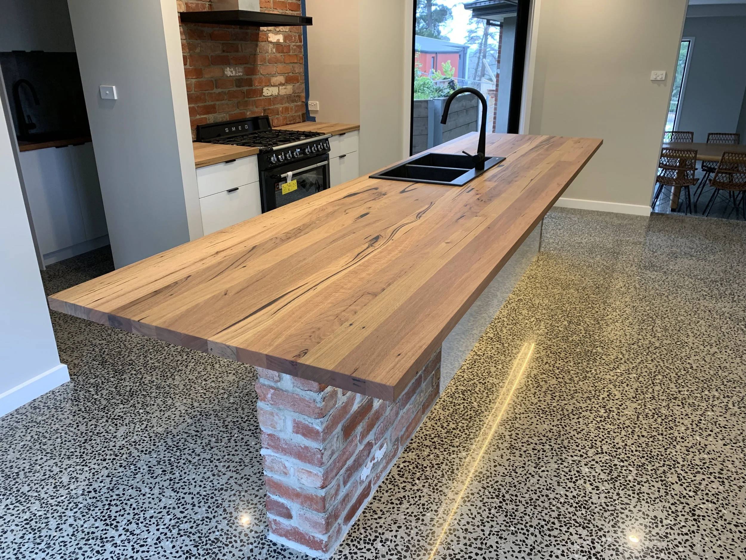 Modern kitchen island with a thick wooden countertop supported by a brick base, black sink and faucet, and a view of a brick wall stove and cabinets behind.