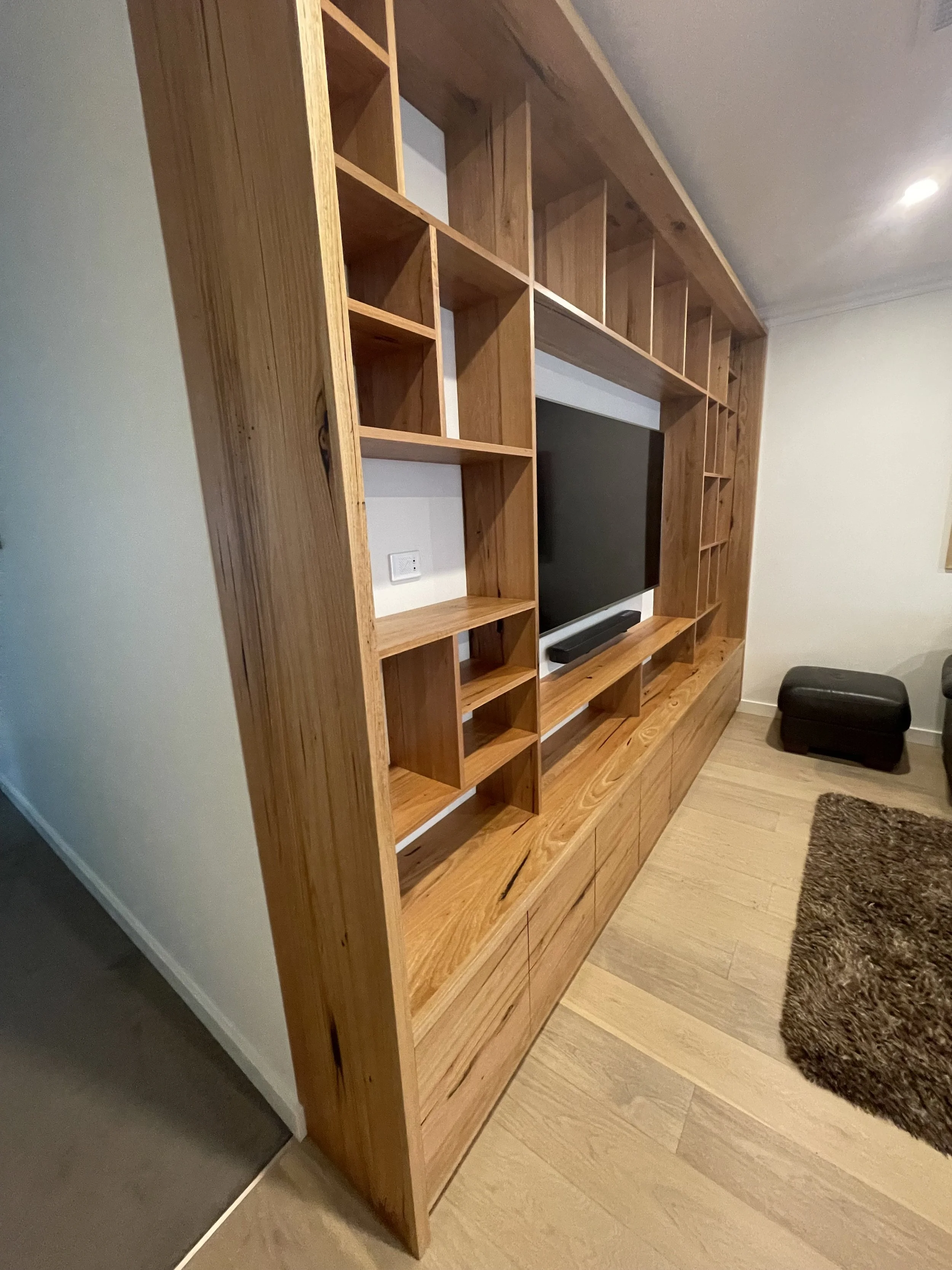Wooden entertainment center with shelves and compartments, holding a flat-screen TV, situated in a living room with a brown rug and black ottoman.