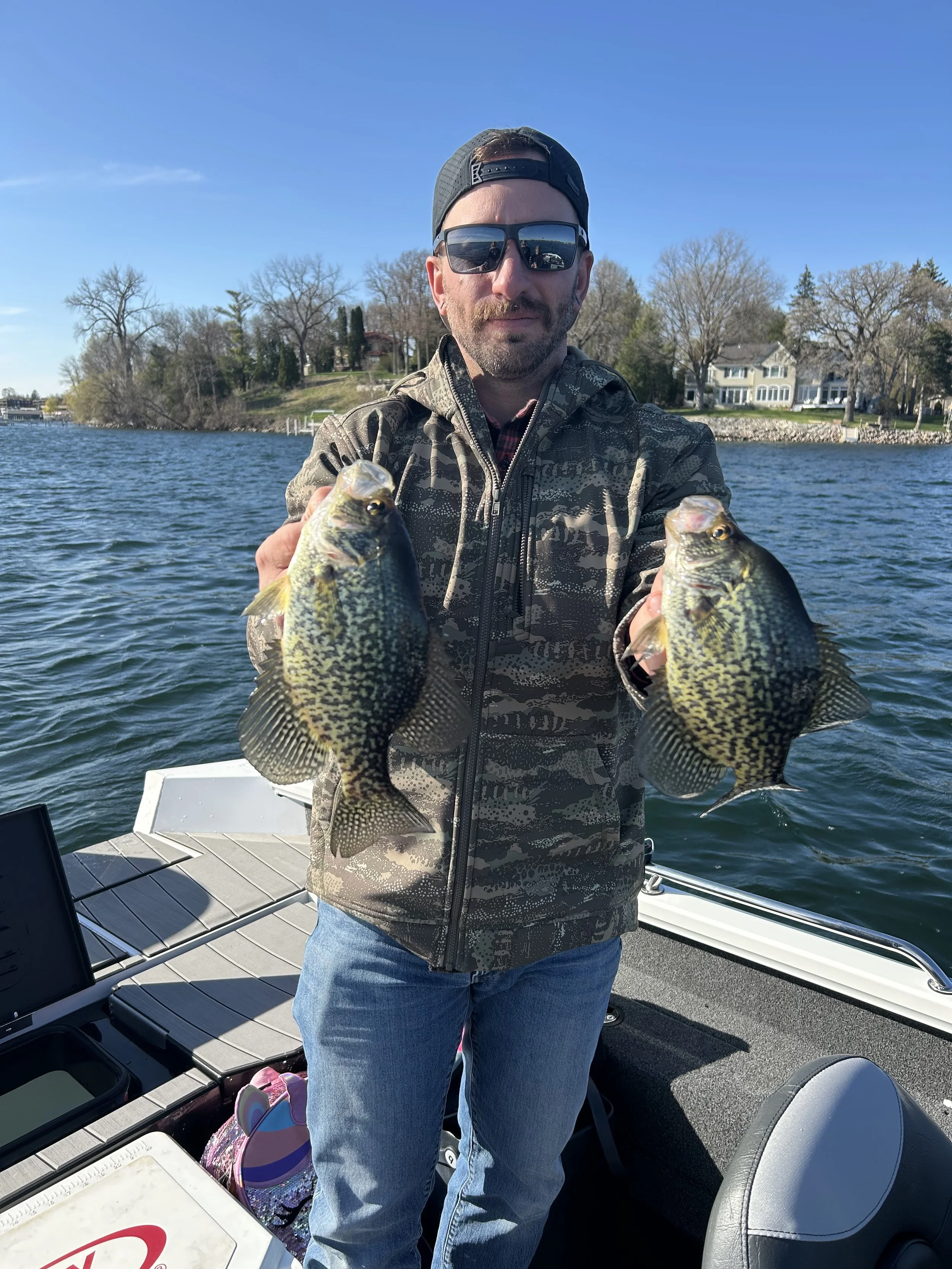 Man on a boat holding two fish, one in each hand, with houses and trees in the background on a sunny day.