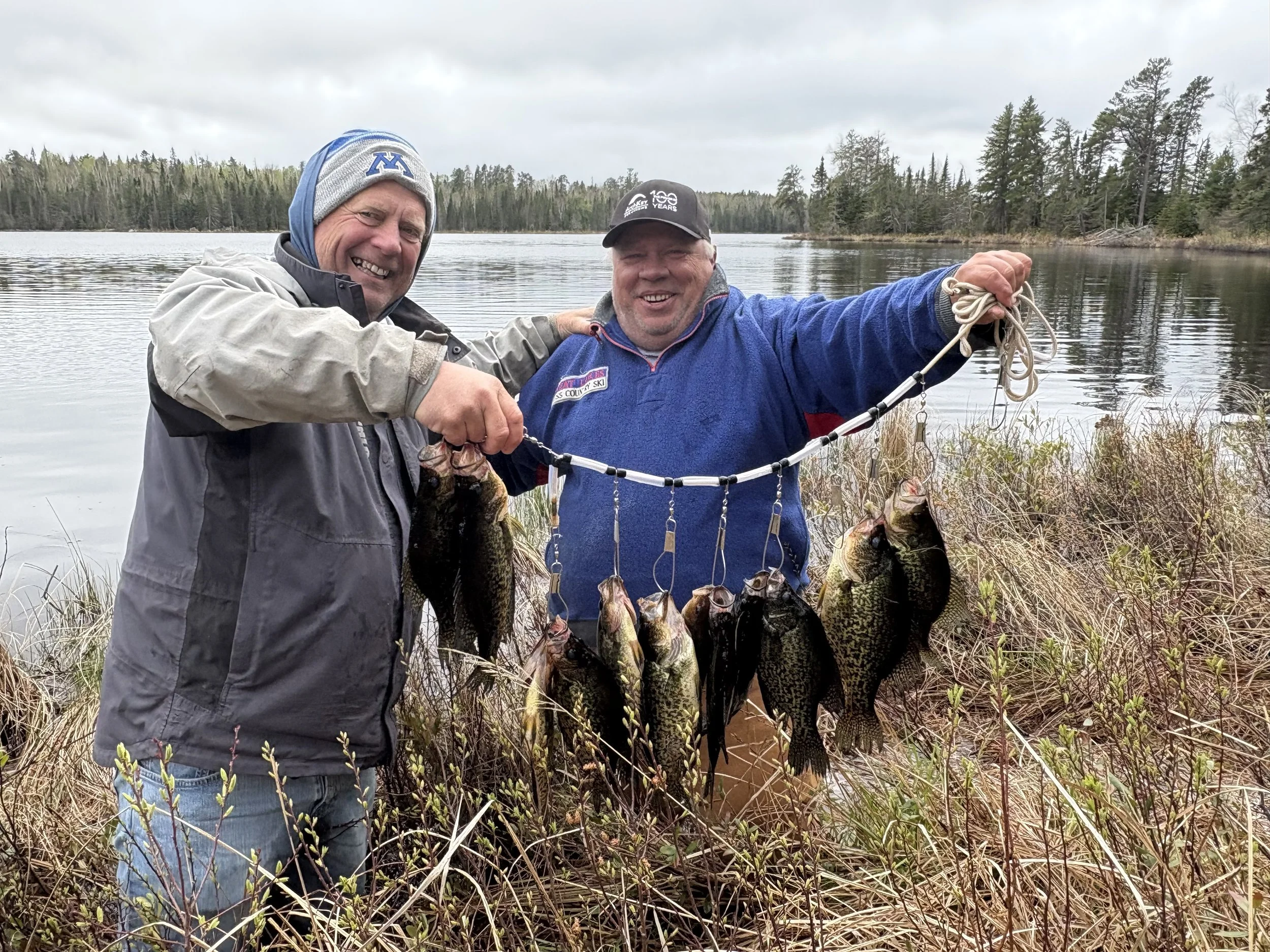 Two men standing by a lakeshore, holding a string of freshly caught fish, smiling and showing their catch, with trees and water in the background.