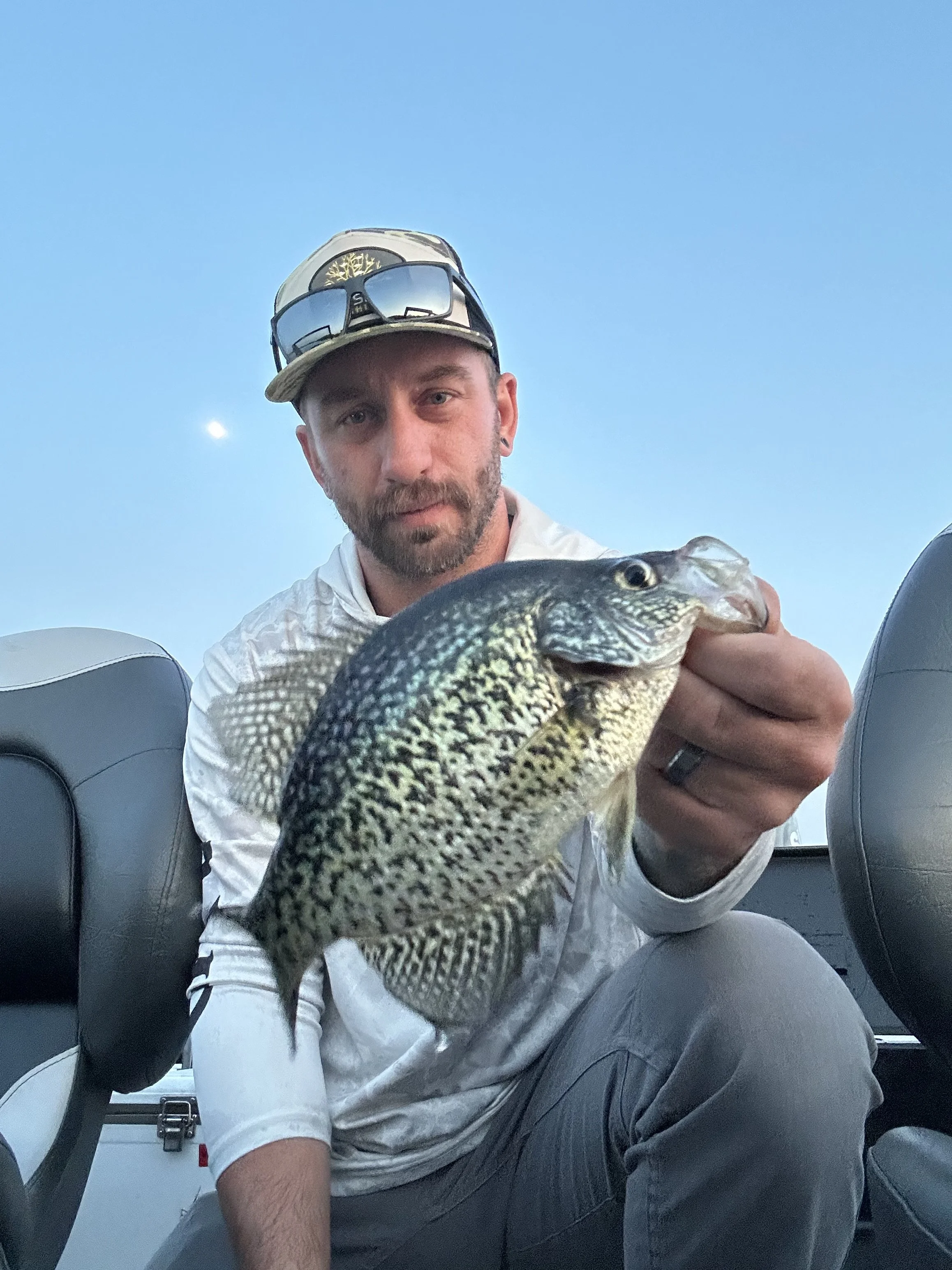 A man on a boat holding a freshly caught fish with a clear blue sky in the background.