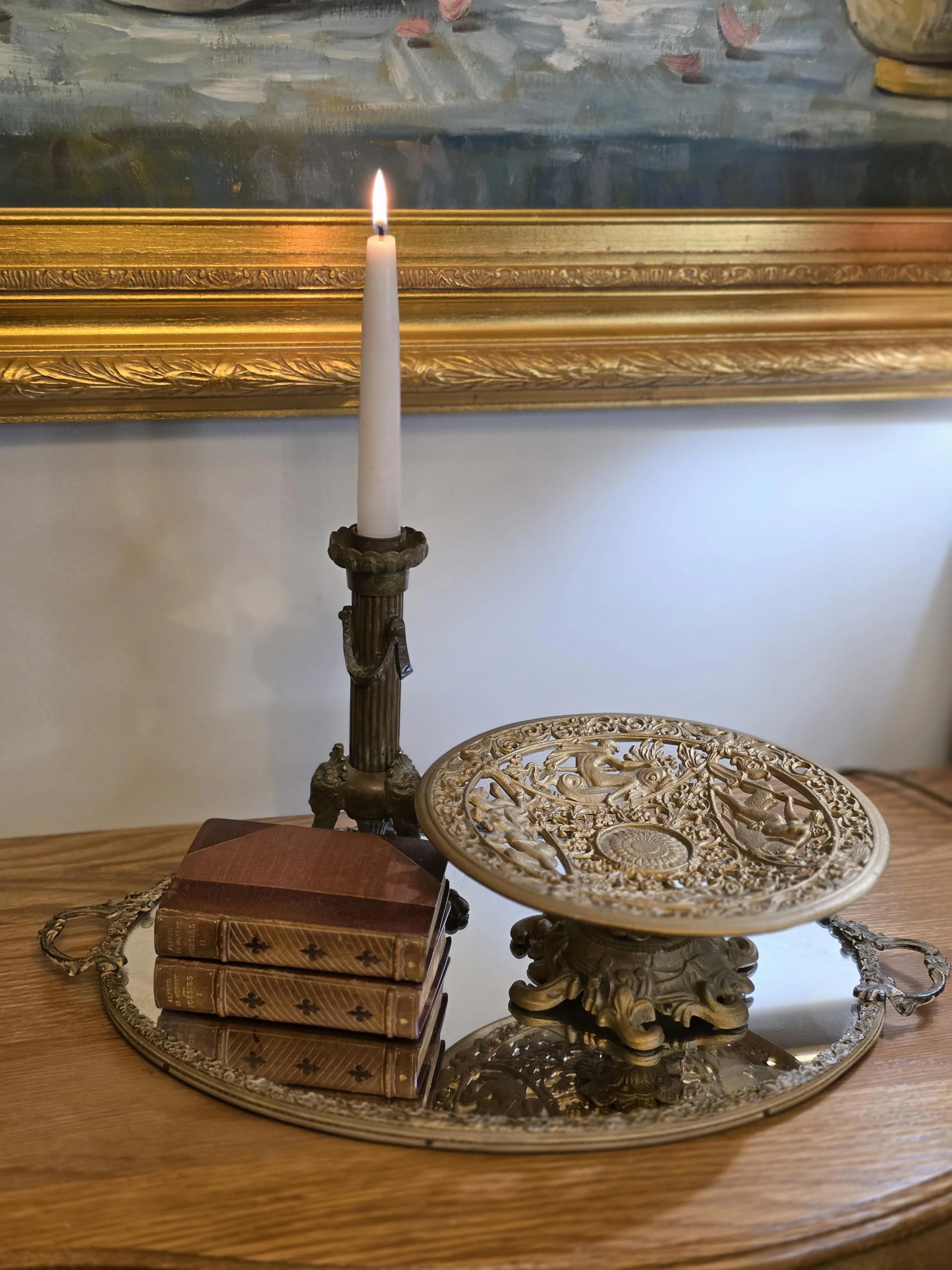 A vintage candle holder with a lit white candle, a stack of three old brown books, a decorative metal plate with intricate designs, all placed on a silver tray on a wooden surface.
