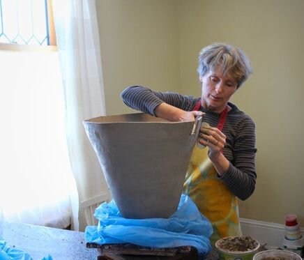 A woman wearing a striped shirt and a yellow apron working with a large clay pot in a bright room.