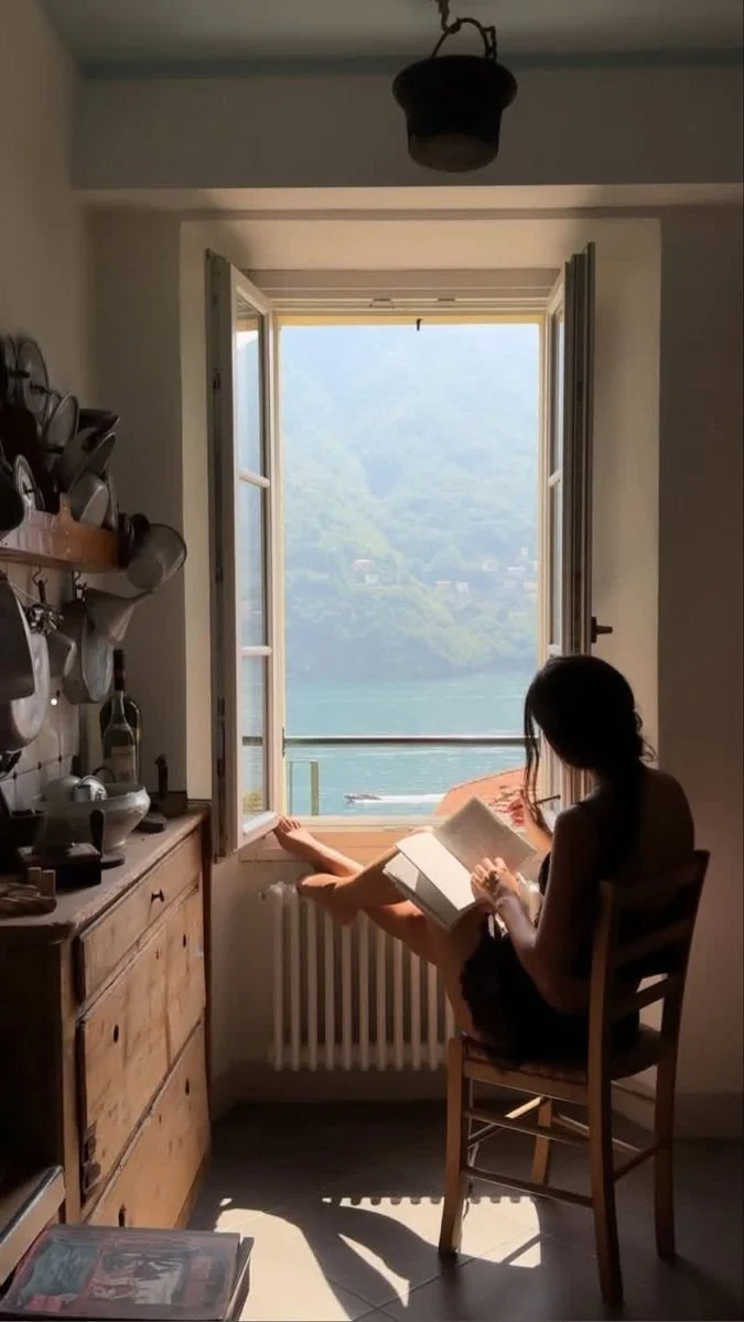 Woman sitting in a chair reading beside a window overlooking a lake and mountains.
