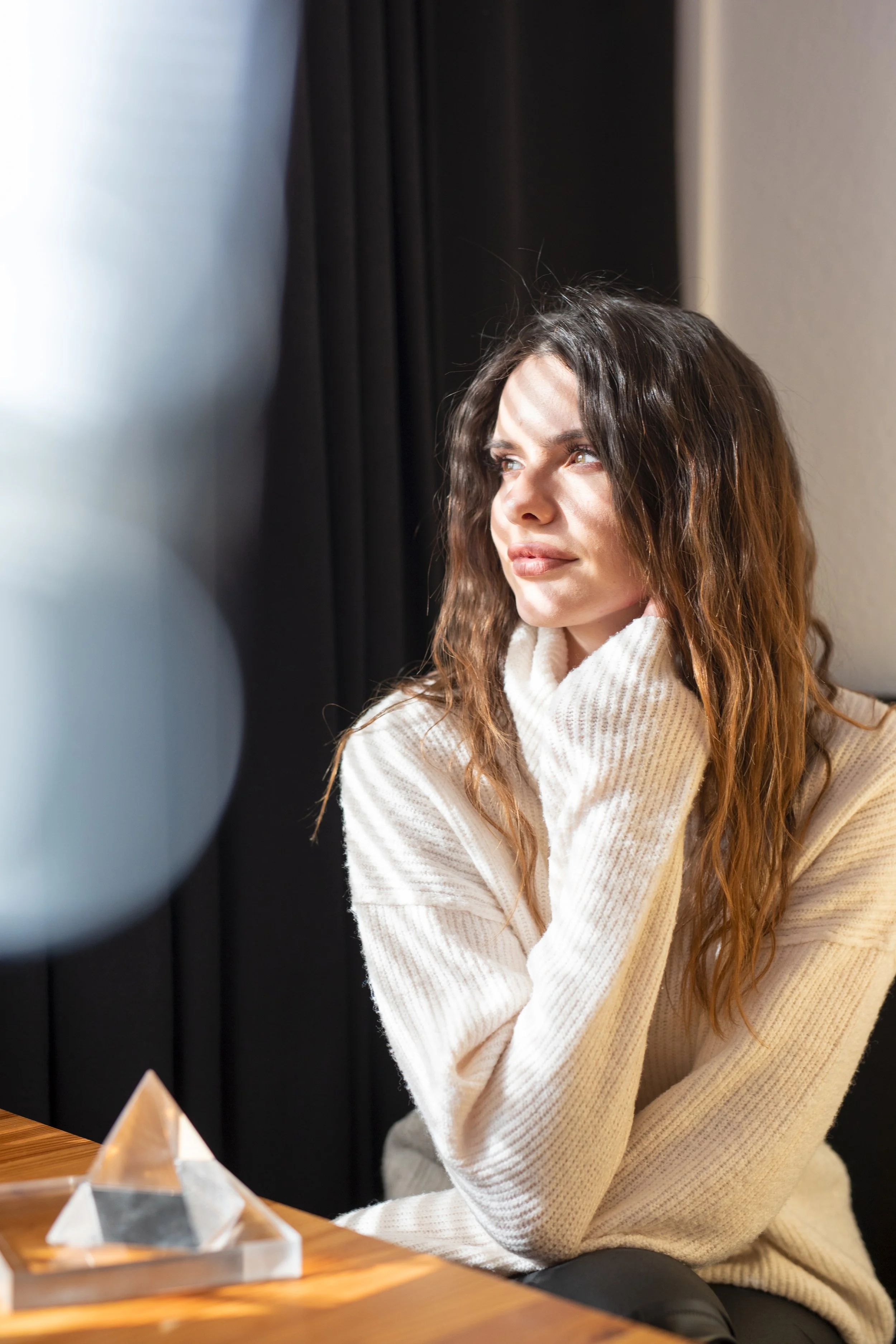 Woman with long wavy brown hair, wearing a cream-colored turtleneck sweater, sitting at a wooden table, looking thoughtfully to the left, with black curtains behind her and a geometric glass sculpture on the table.