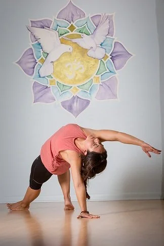 Woman practicing yoga in a peace sign pose in front of a mural of two white doves, a moon, and a lotus flower on a light-colored wall.