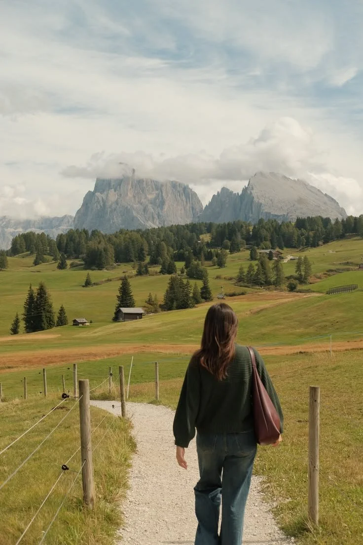 Woman walking along a gravel path through a green landscape with mountains in the background.