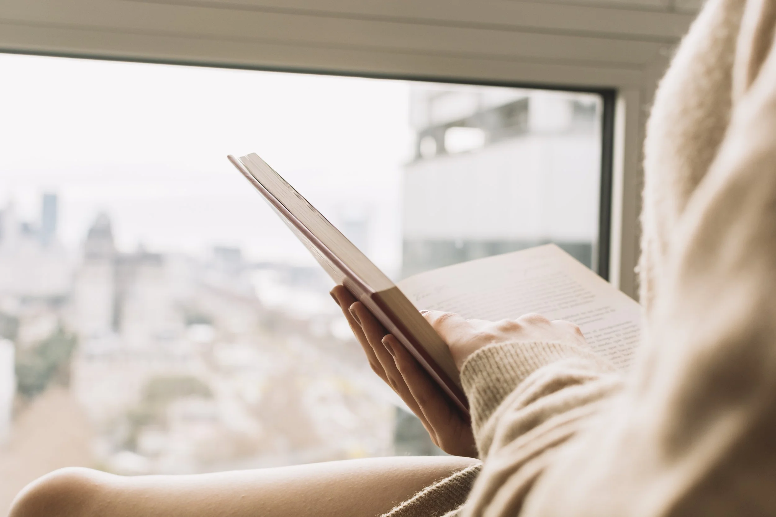 Person sitting by a window reading a book, with a cityscape view outside.