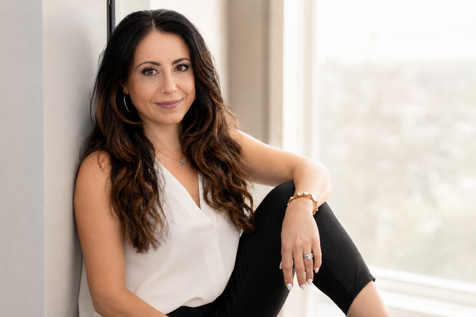 A woman with long, wavy dark hair wearing a white sleeveless top, black pants, sitting near a window, smiling, with jewelry including hoops, a necklace, bracelets, and a ring.
