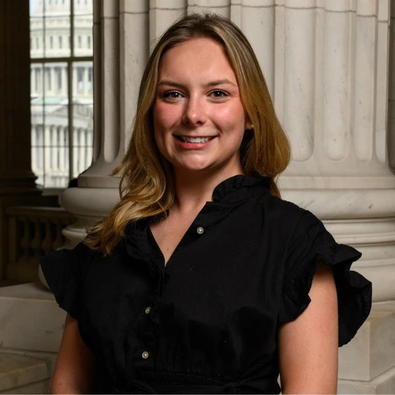 A young woman with shoulder-length blonde hair, wearing a black blouse with ruffled sleeves, smiling and standing in front of a classical architectural column inside a building with large windows.