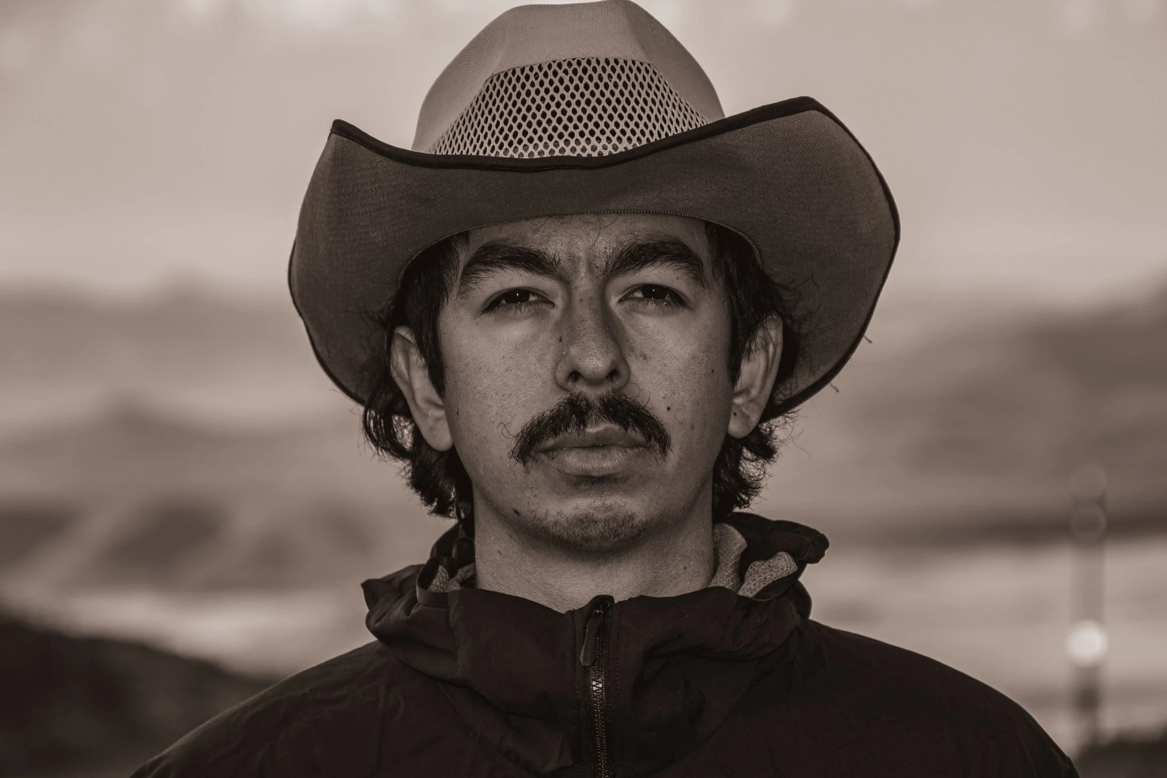A man wearing a cowboy hat and a jacket, looking directly at the camera with a serious expression, outdoors in a desolate landscape.