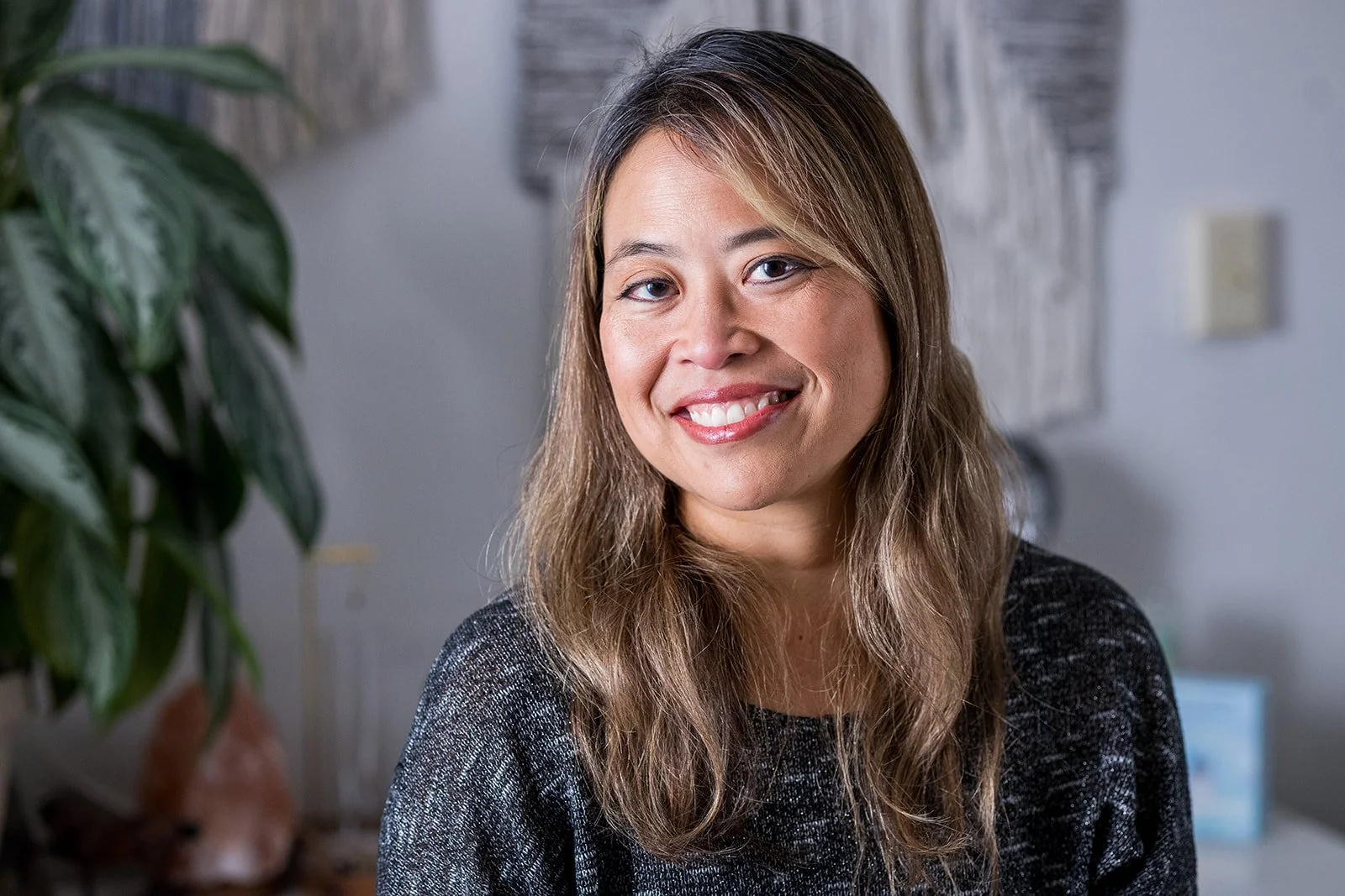 A woman with long wavy light brown hair, wearing a black top, smiling in a room with a plant and wall decorations.