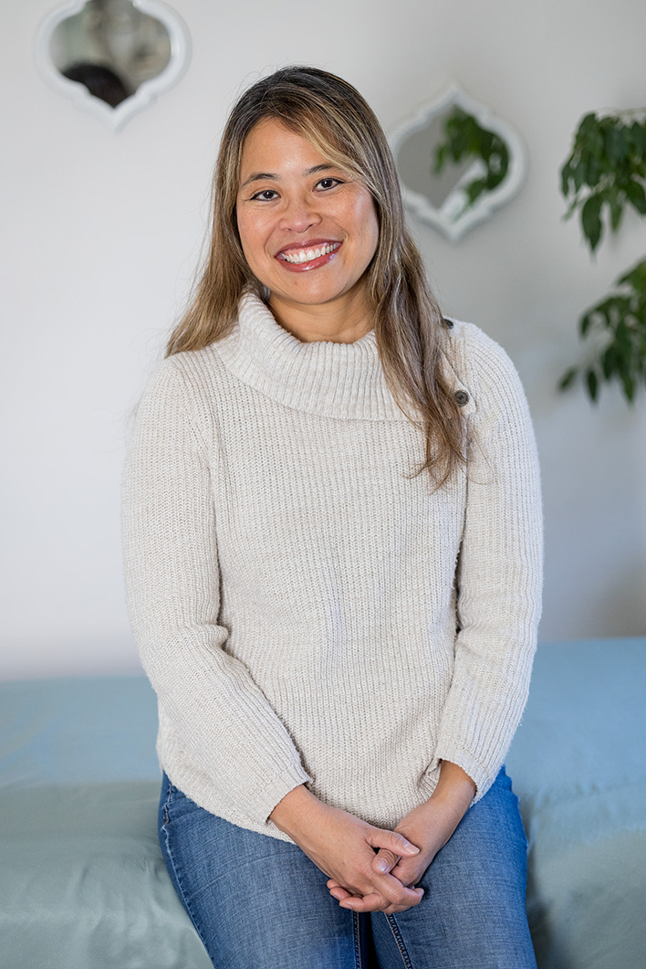A woman with long light brown hair and a bright smile, wearing a cream-colored knit sweater and blue jeans, sitting in a cozy room with a bed and decorative wall mirrors in the background.
