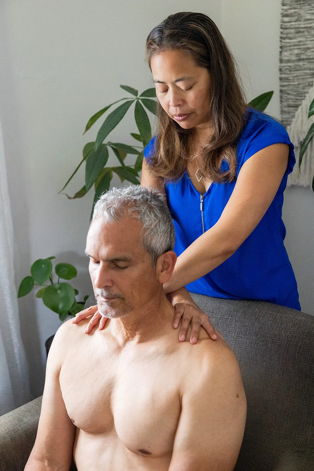 Shift Mind Body Spirit_Myofascial Release Therapy_Berkeley, CA_A woman giving a massage to a shirtless man on a grey couch in a room with green plants.