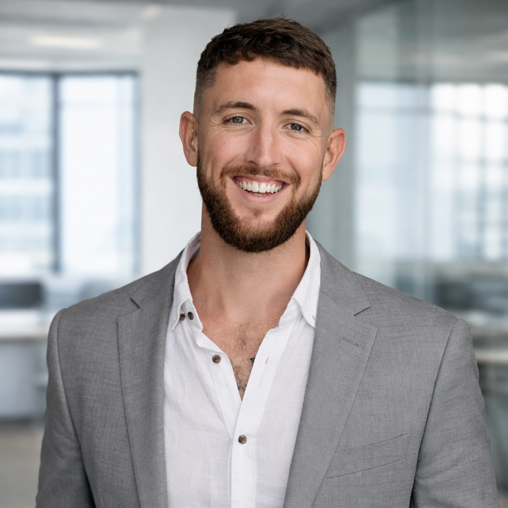 A young man with a beard and short brown hair smiling, wearing a light gray suit jacket over a white shirt, standing in an office setting.