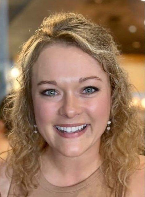 A woman smiling with curly blonde hair, wearing earrings and a light-colored top, indoors with warm lighting.