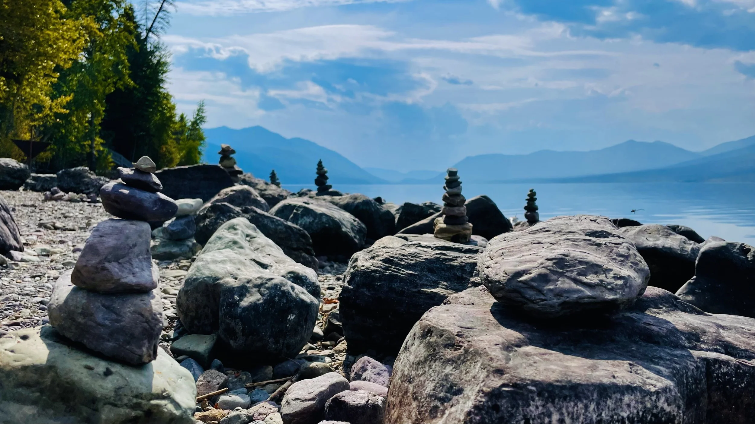 A rocky lakeshore with several small rock stacks, green trees on the left, mountains in the background, and a partly cloudy blue sky.