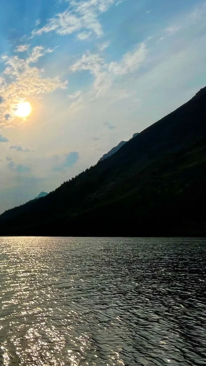 Scenic photo of a mountain lake during sunset, with the sun partially obscured by clouds, mountain silhouette on the right, and water reflecting the sky.