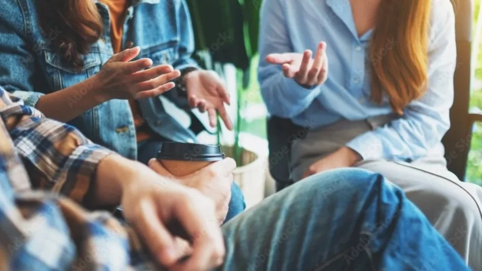 Three people are having a conversation, with one person holding a coffee cup and gesturing with their hands, while the others listen and also gesture, in a cozy, natural light setting.