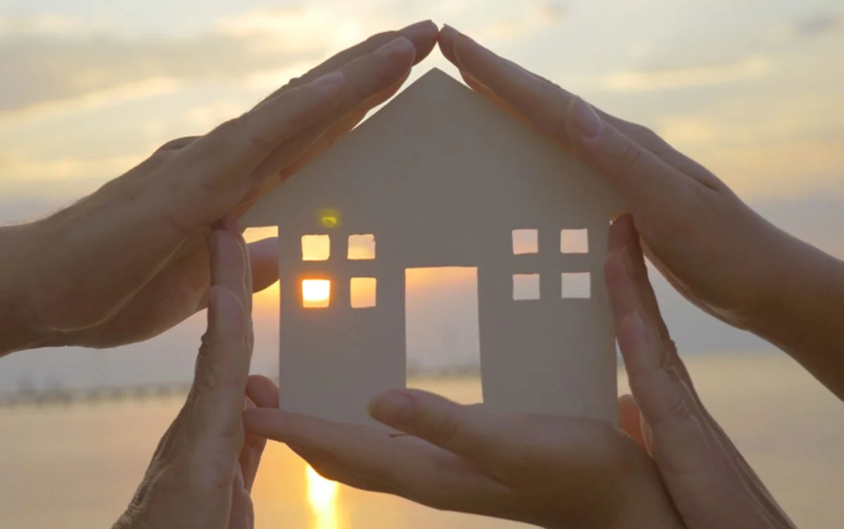 Hands holding a paper house cutout against a sunset background with the sun shining through windows.