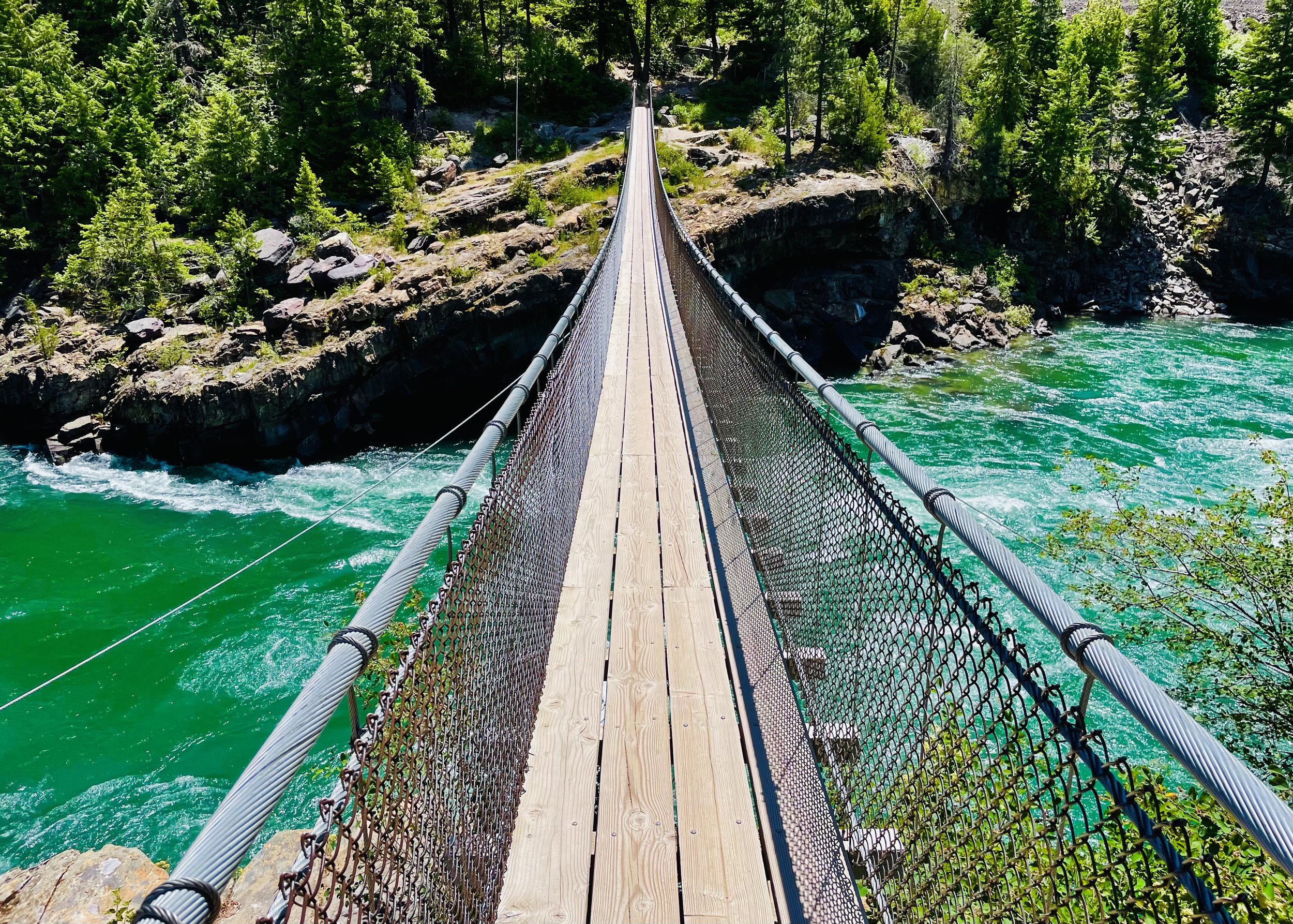 A wooden suspension bridge with metal railings spans over a river with turquoise water, surrounded by lush green trees and rocky terrain.