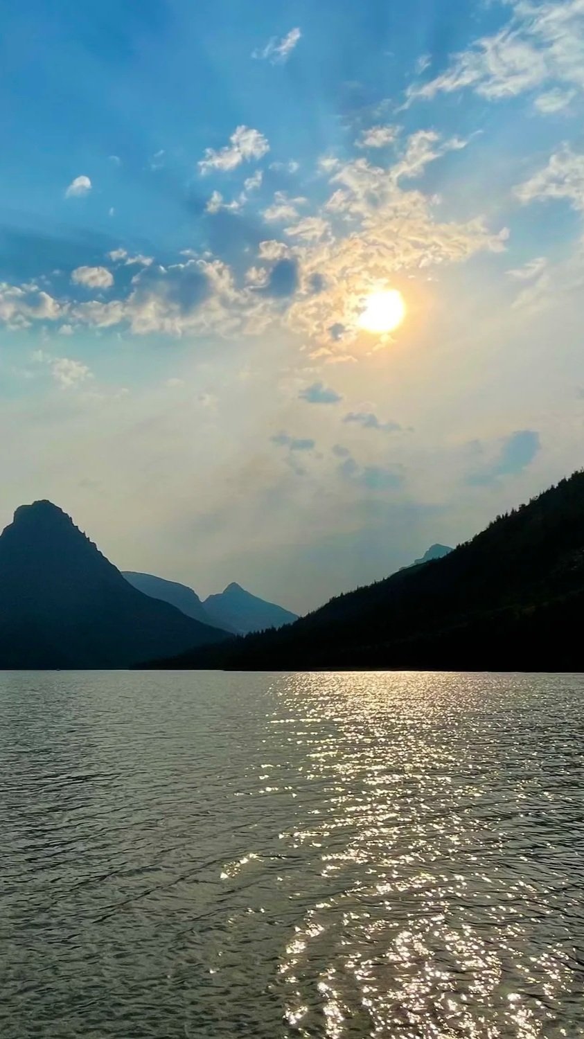 Sunset over a lake surrounded by mountains, with partly cloudy sky and shimmering water reflections.
