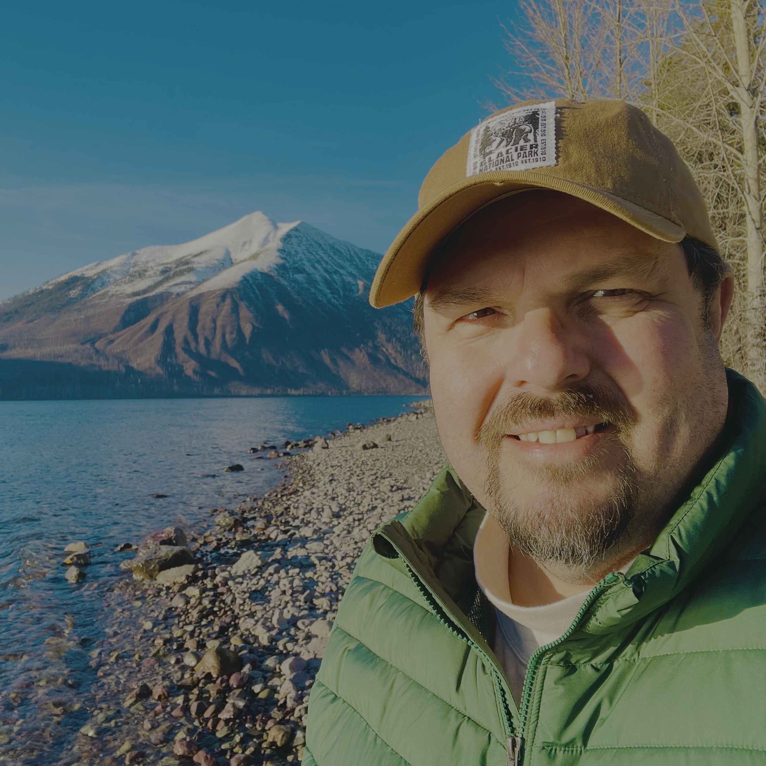 Close-up of a smiling man's face wearing a brown and green cap, with a mountain and lake landscape in the background.