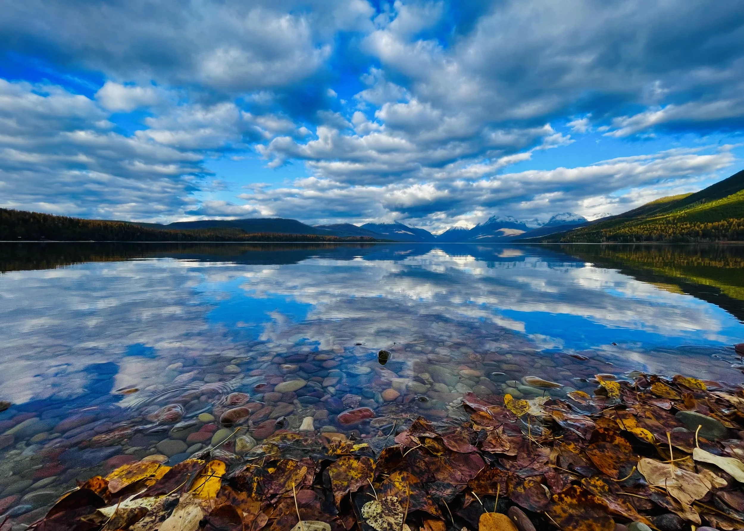 Scenic view of a calm mountain lake with clear water reflecting the blue sky and white clouds, surrounded by hills with green trees and distant snow-capped mountains.