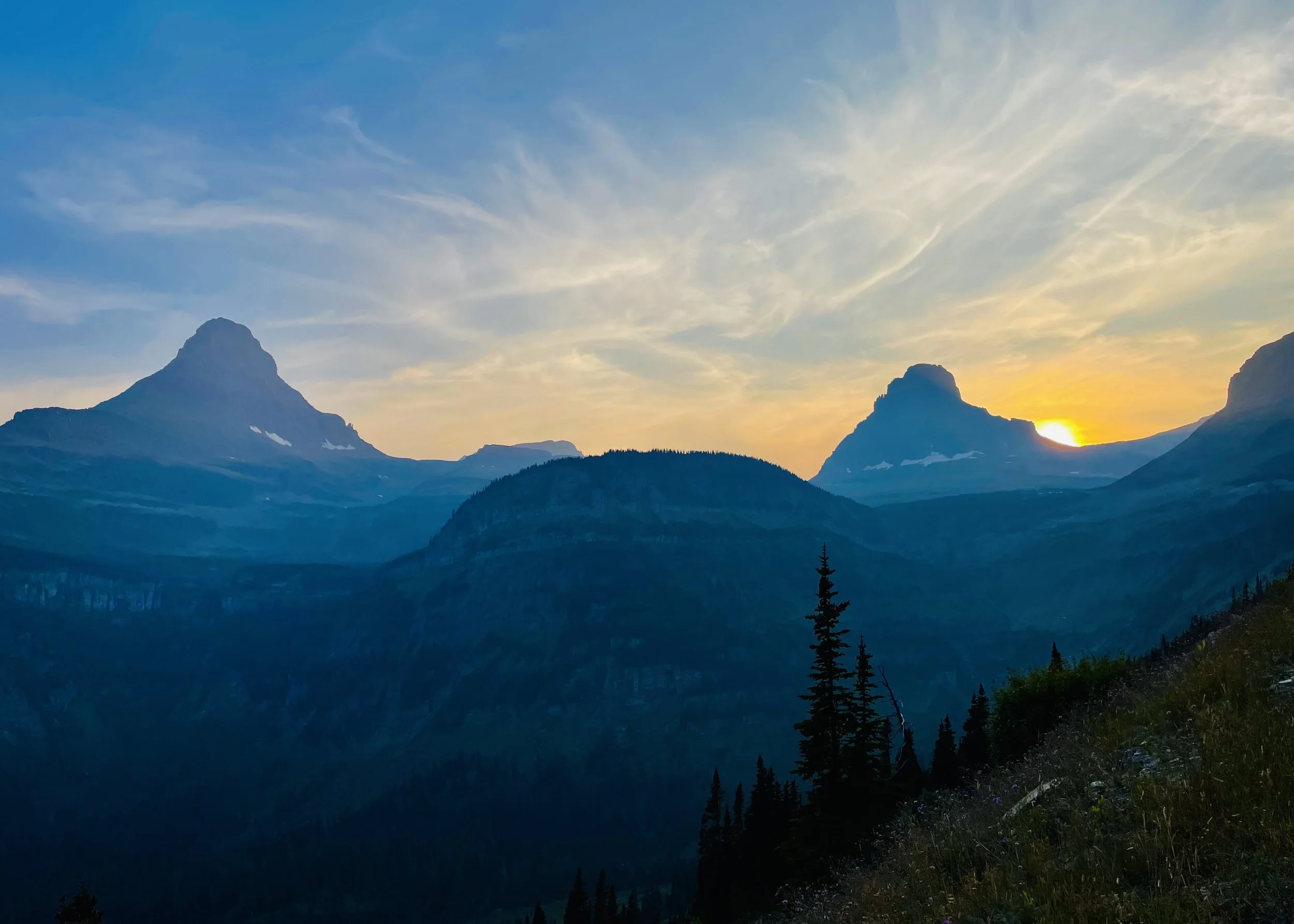 Mountain landscape at sunset with peaks, forests, and a partly cloudy sky.