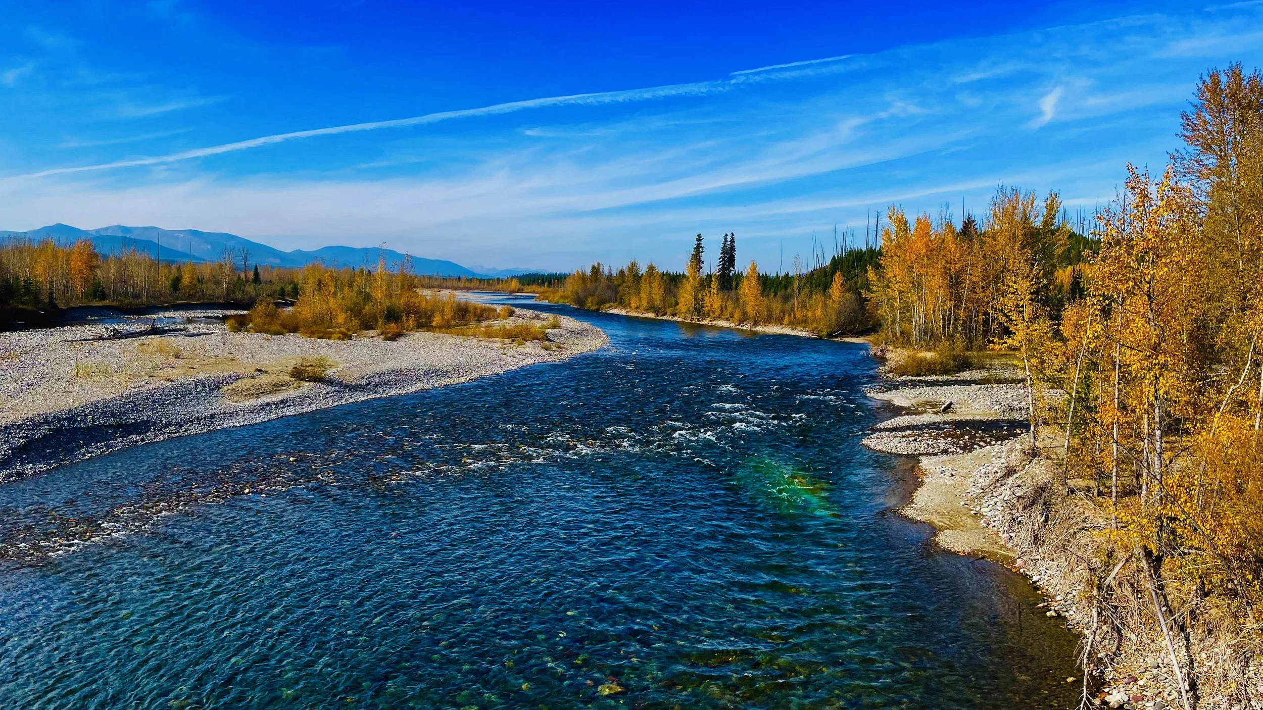 A river flowing through a landscape with colorful autumn trees on the banks and mountains in the distance under a blue sky with wispy clouds.